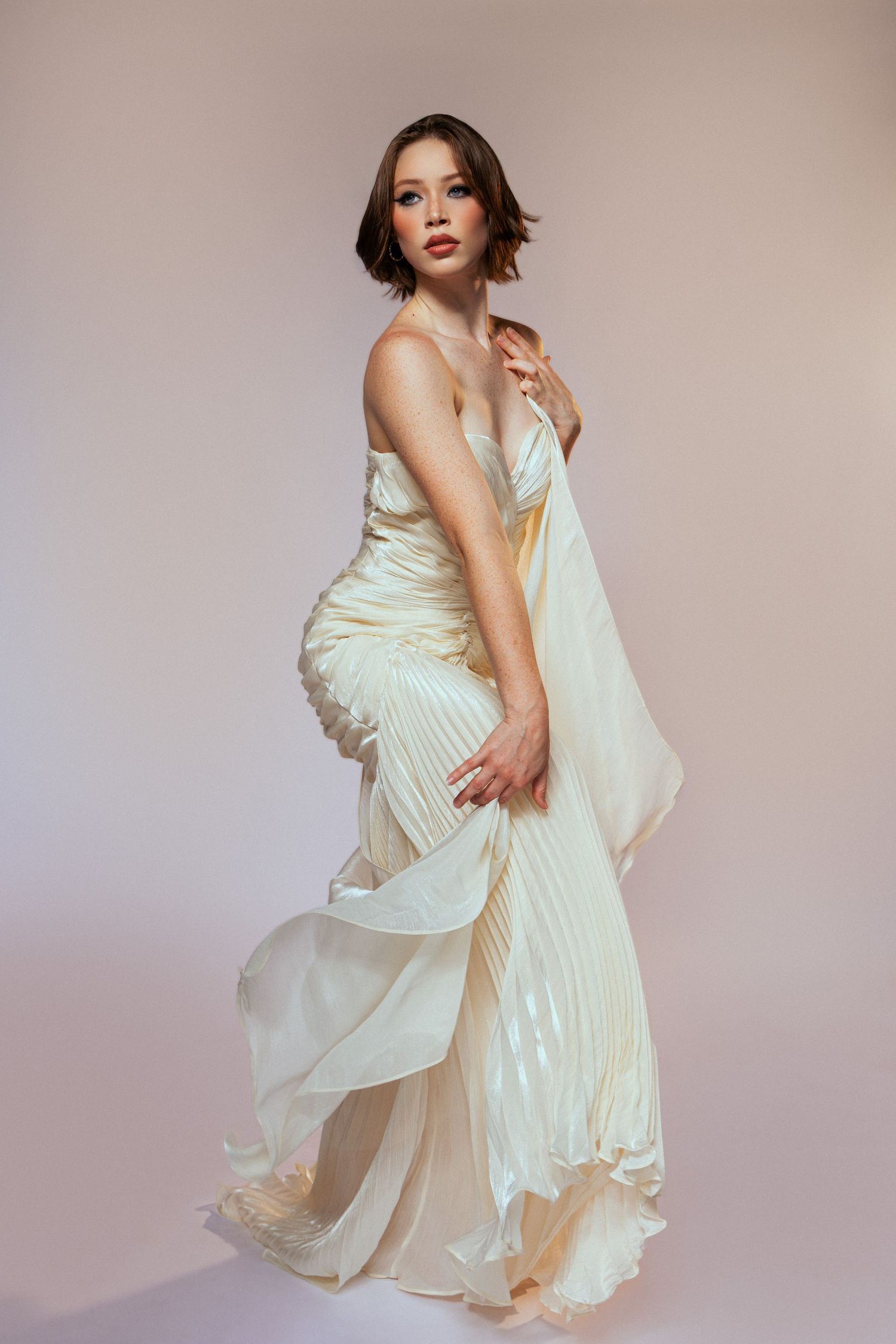 Fashion model poses gracefully in a flowing white pleated dress against a neutral studio backdrop.