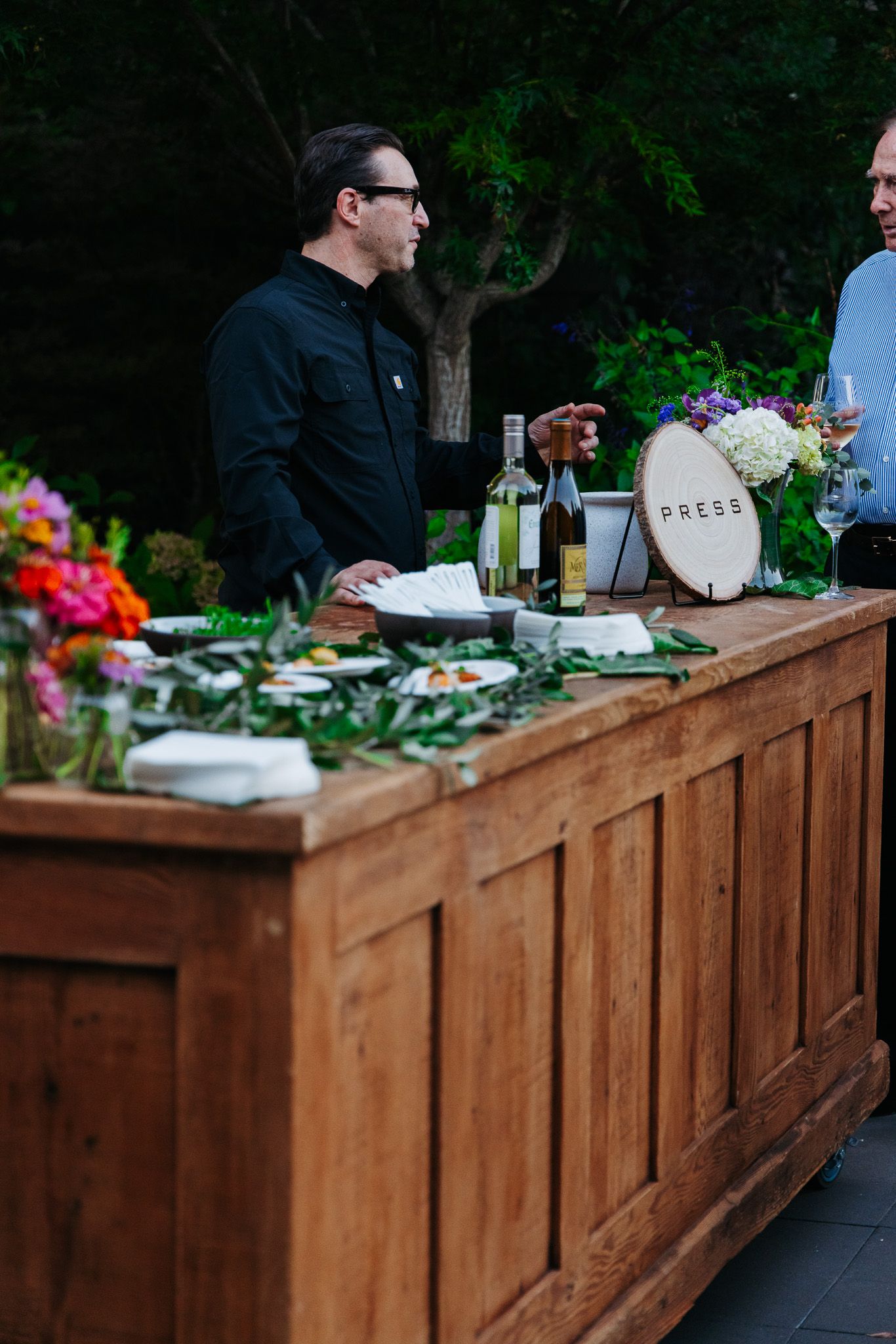 People serve themselves food from a rustic wooden buffet station decorated with colorful flowers at an outdoor event.