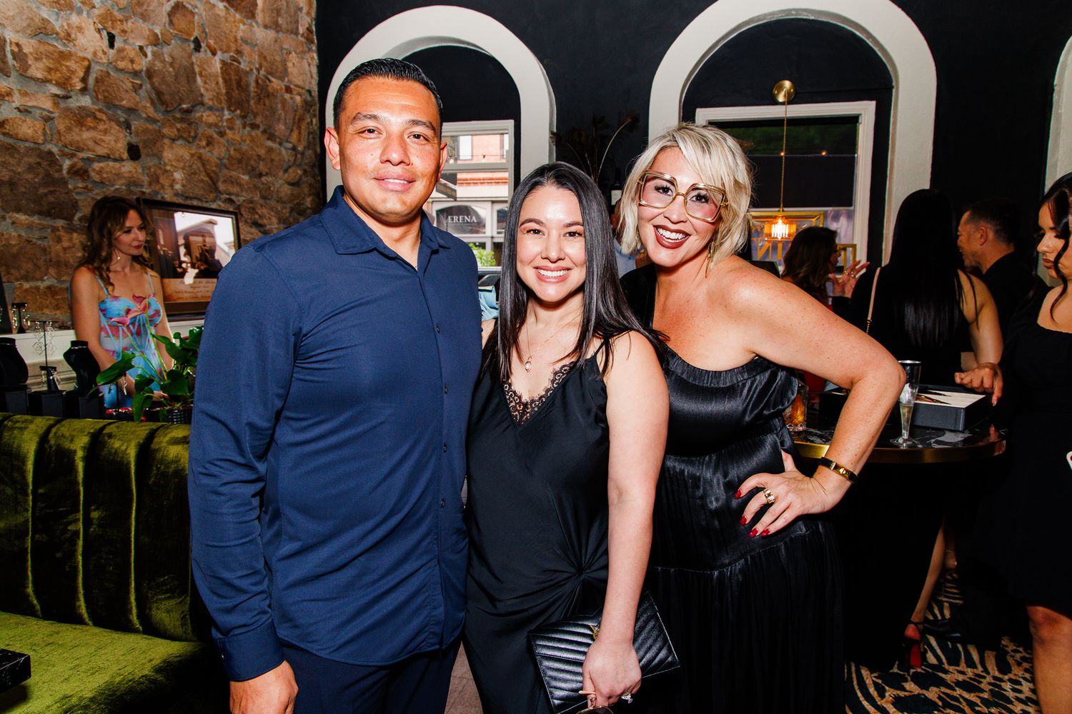 Three friends smile together at an upscale evening event in a stylish venue with green velvet seating.