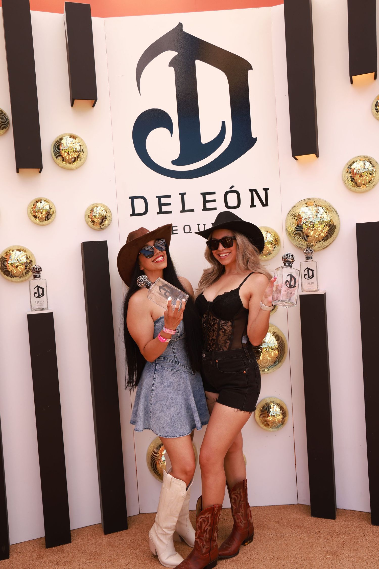 Two women in cowboy hats pose at a DeLeon Tequila branded backdrop with gold disco balls.