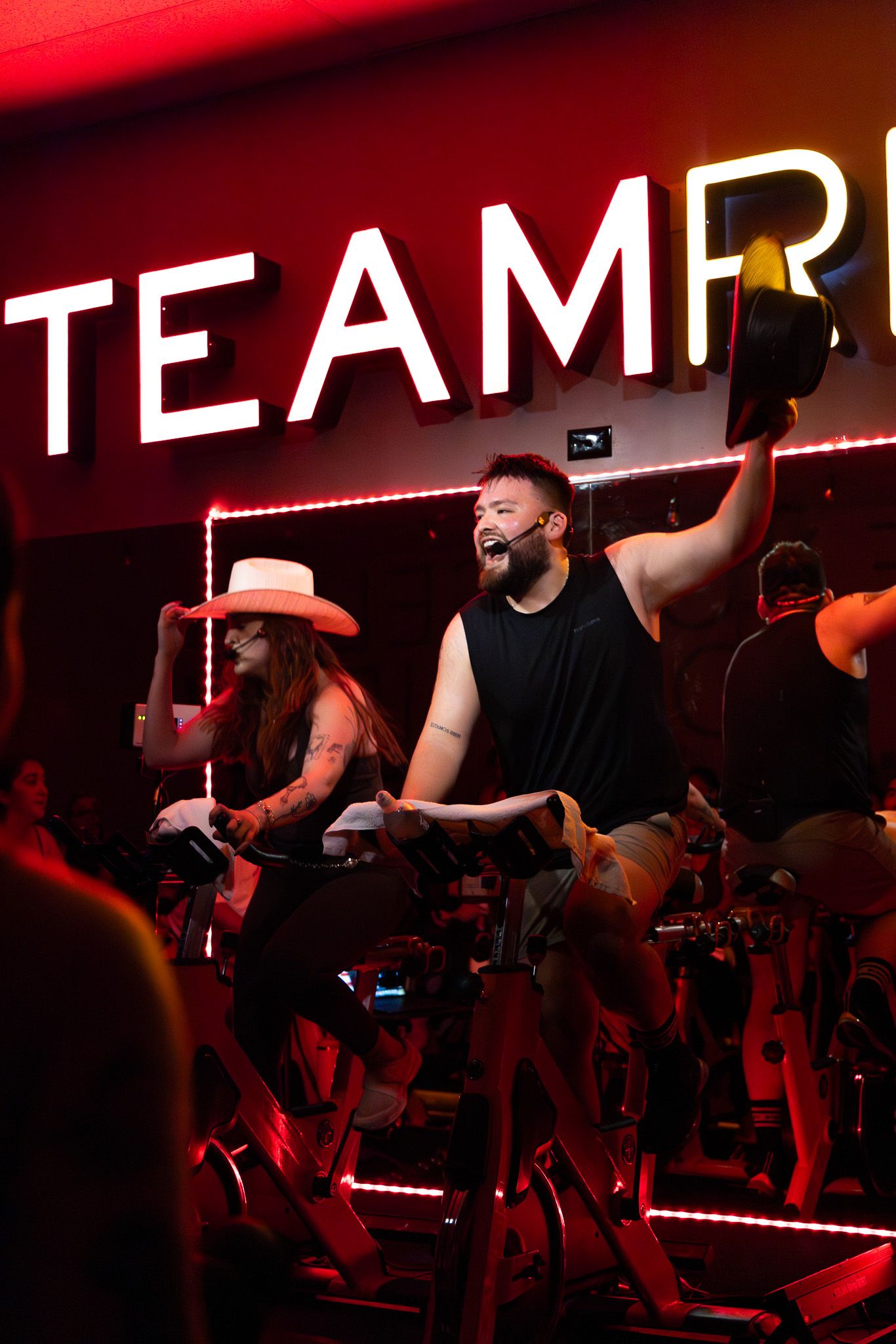 An energetic spin class instructor leads a workout session under red neon TEAMP signage.
