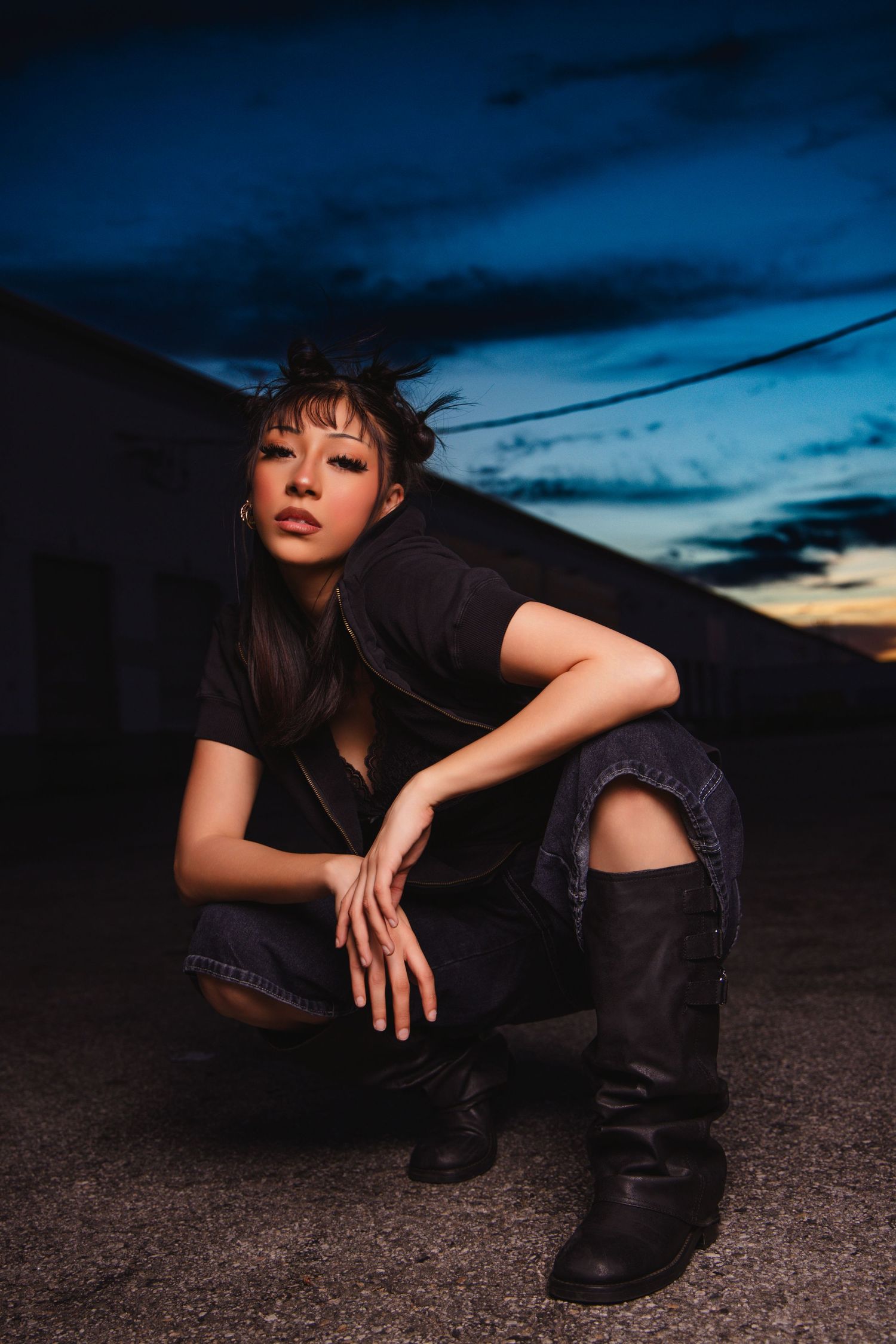 Person in black outfit and ripped jeans crouches on ground against dramatic sunset sky with dark clouds.