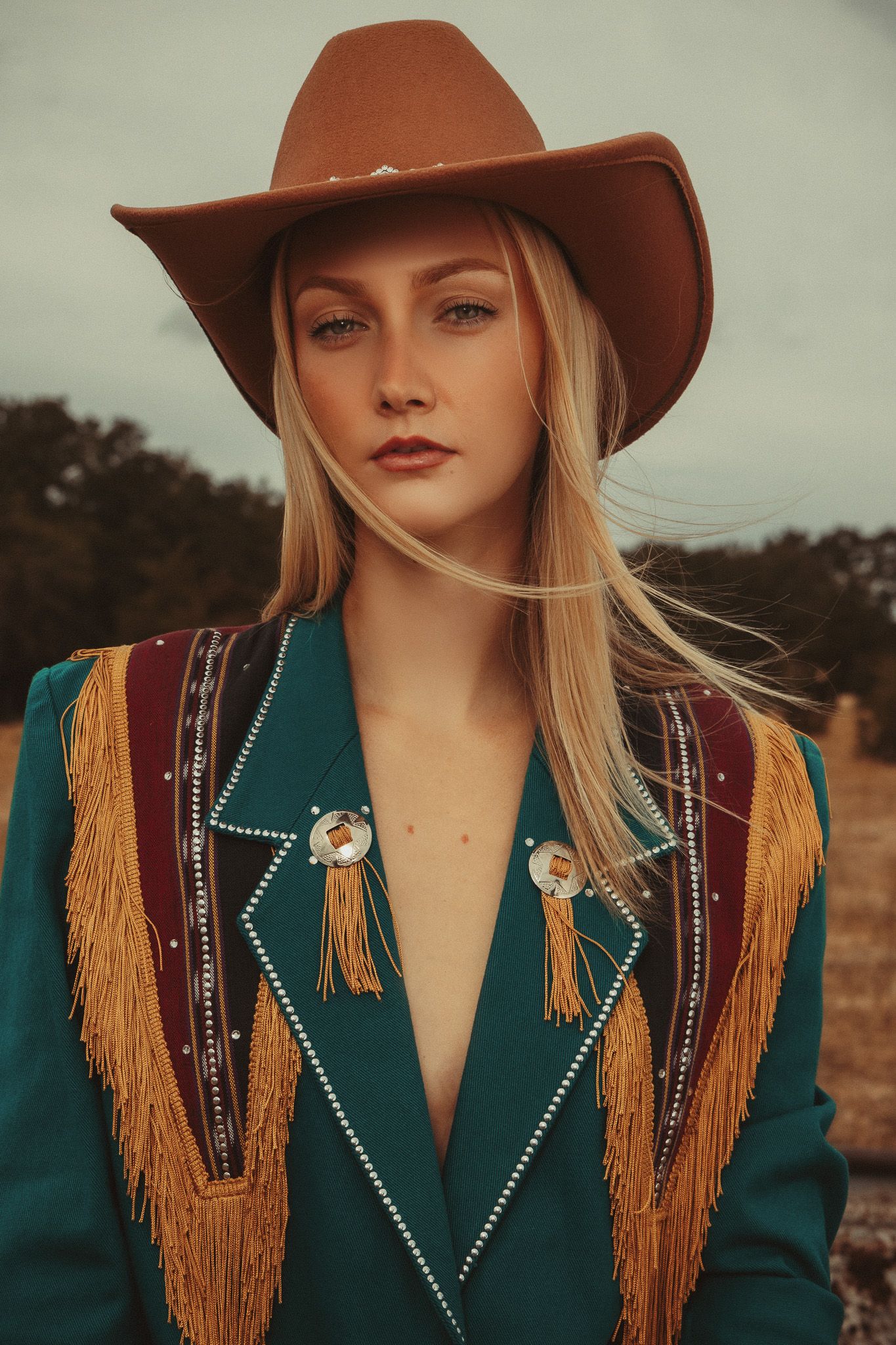 A western-style outfit featuring a teal blazer with gold fringe trim, brown cowboy hat, and studded details against a moody sky.