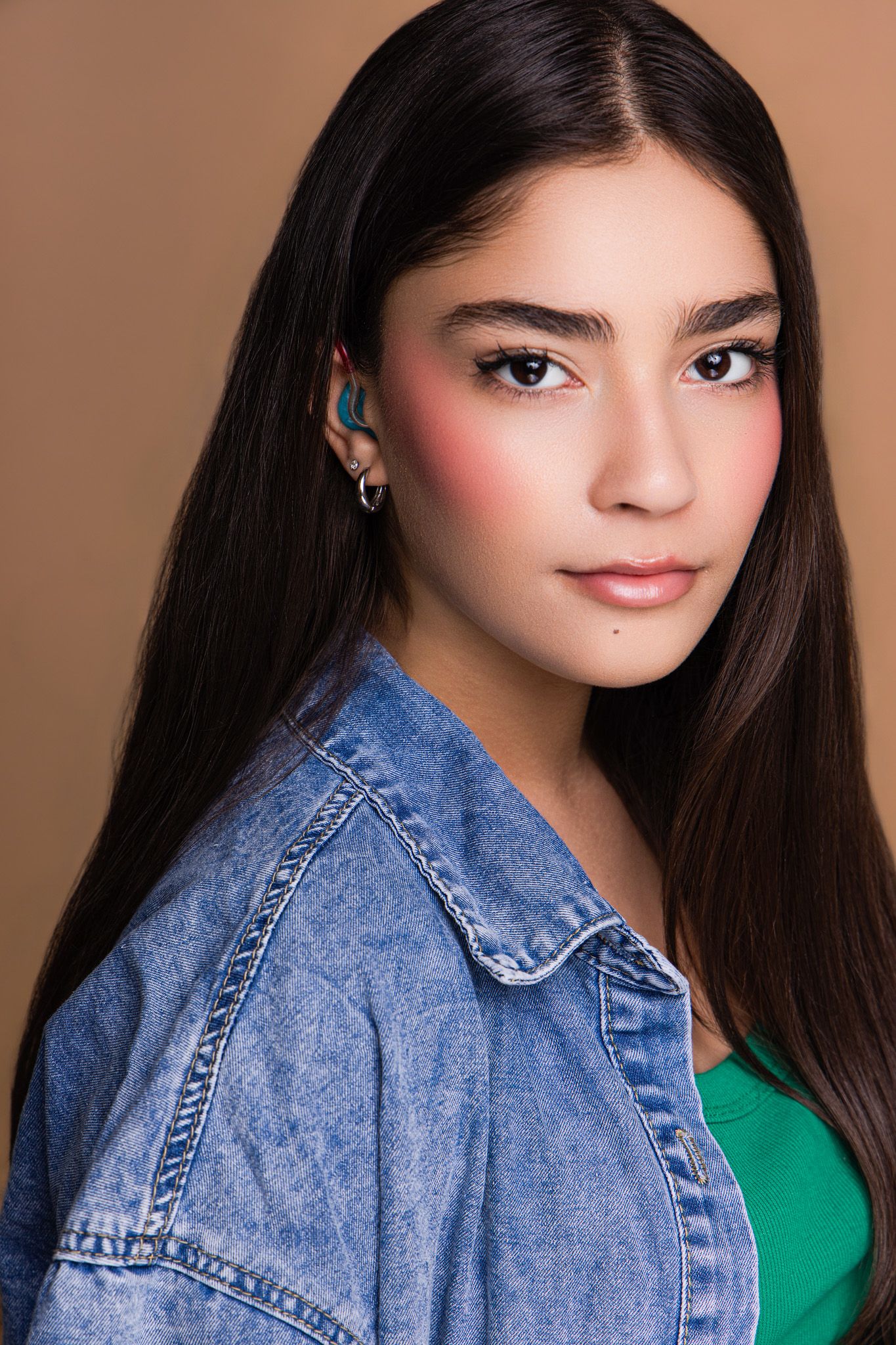 Young woman with long dark hair, rosy blush makeup, small earring, wearing a denim jacket over a green top.