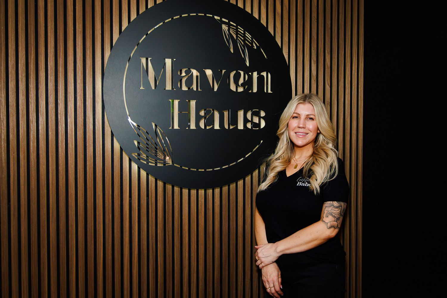 Woman posing in front of Maven Haus circular logo sign on wooden slat wall.