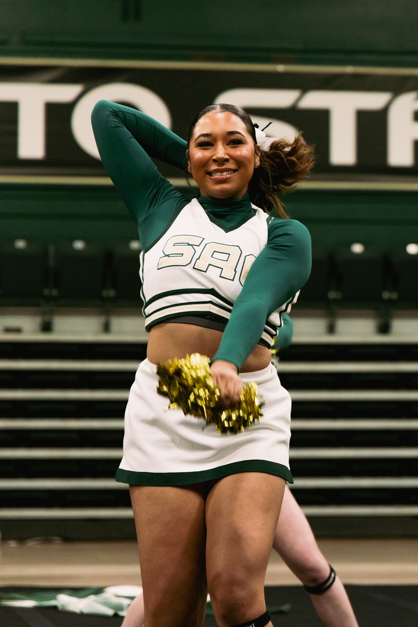 Sacramento State cheerleader smiles while performing with gold pom-poms in green and white SAC uniform.