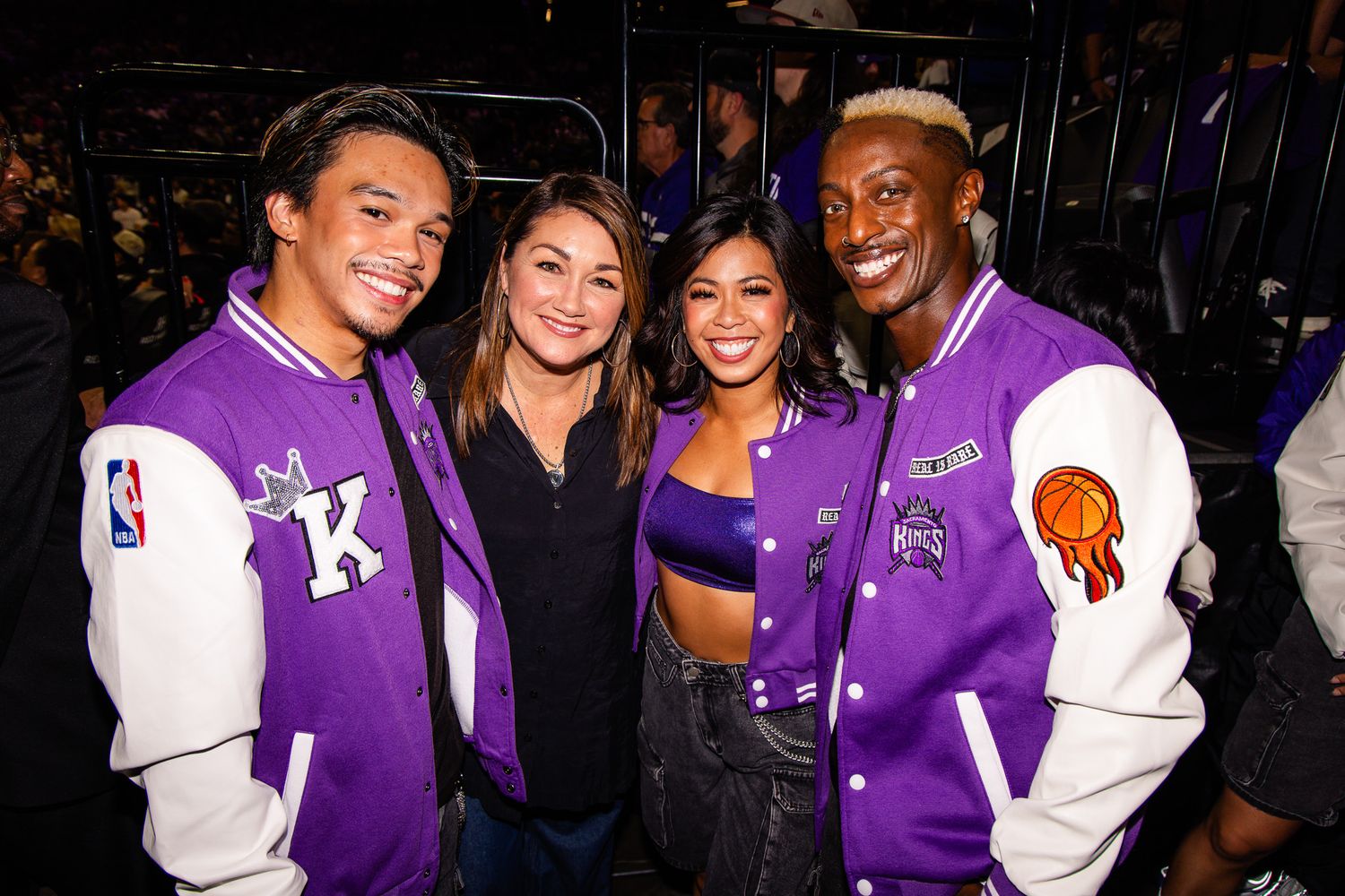 Four friends pose together at an NBA event, two wearing purple and white varsity jackets with the Kings logo.
