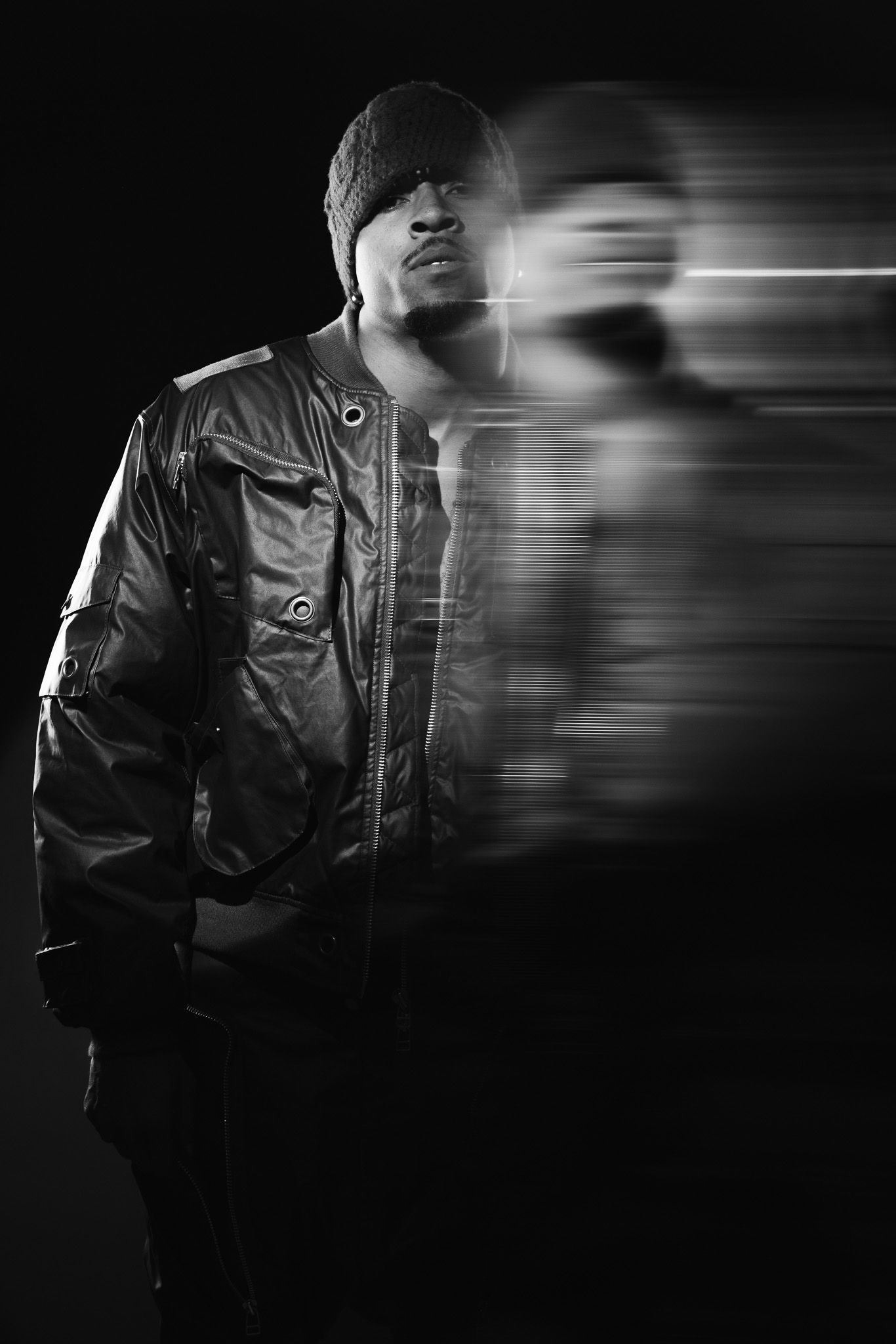 Black and white portrait of a man in a bomber jacket and beanie with a dramatic motion blur effect on dark background.