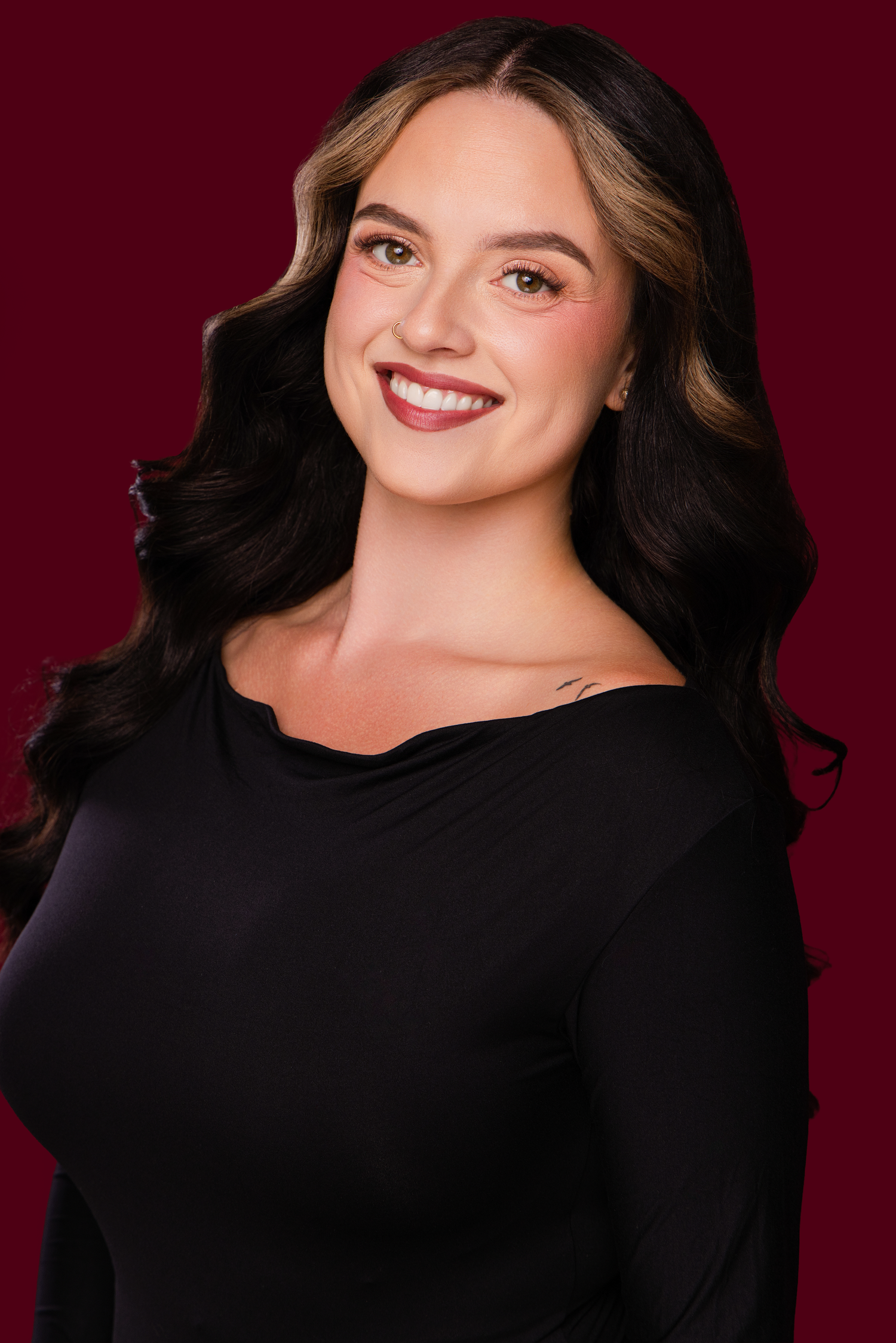 Smiling young woman with long dark wavy hair wearing a black off-shoulder top against a deep red background.