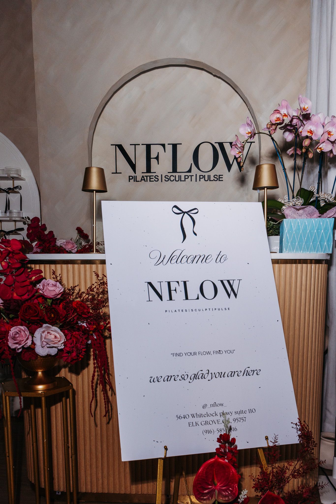 A welcome sign for NFLOW boutique displayed with pink and red floral decorations and gold accent lamps.
