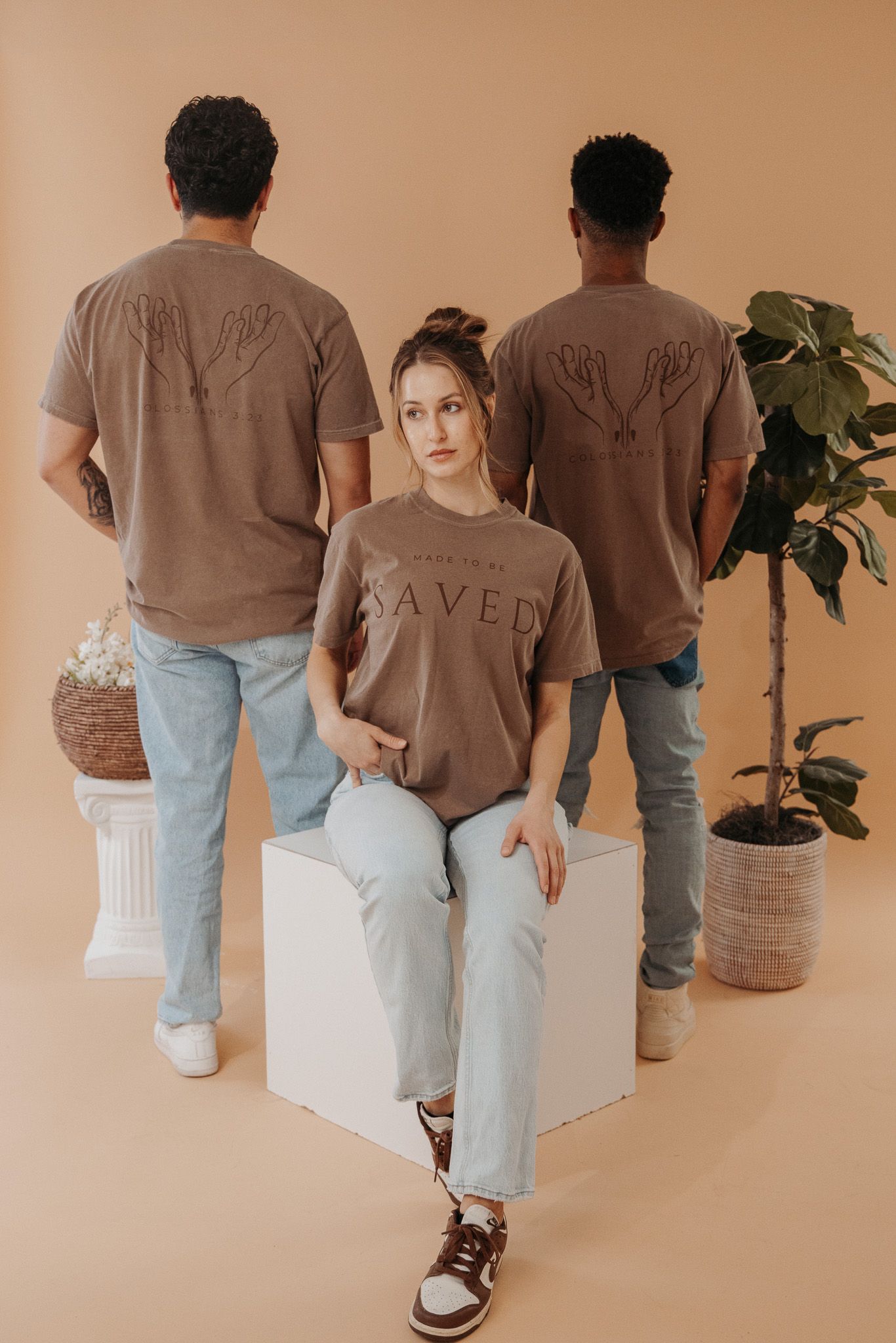 A line of people wearing brown t-shirts and light denim jeans photographed from behind against a beige wall.