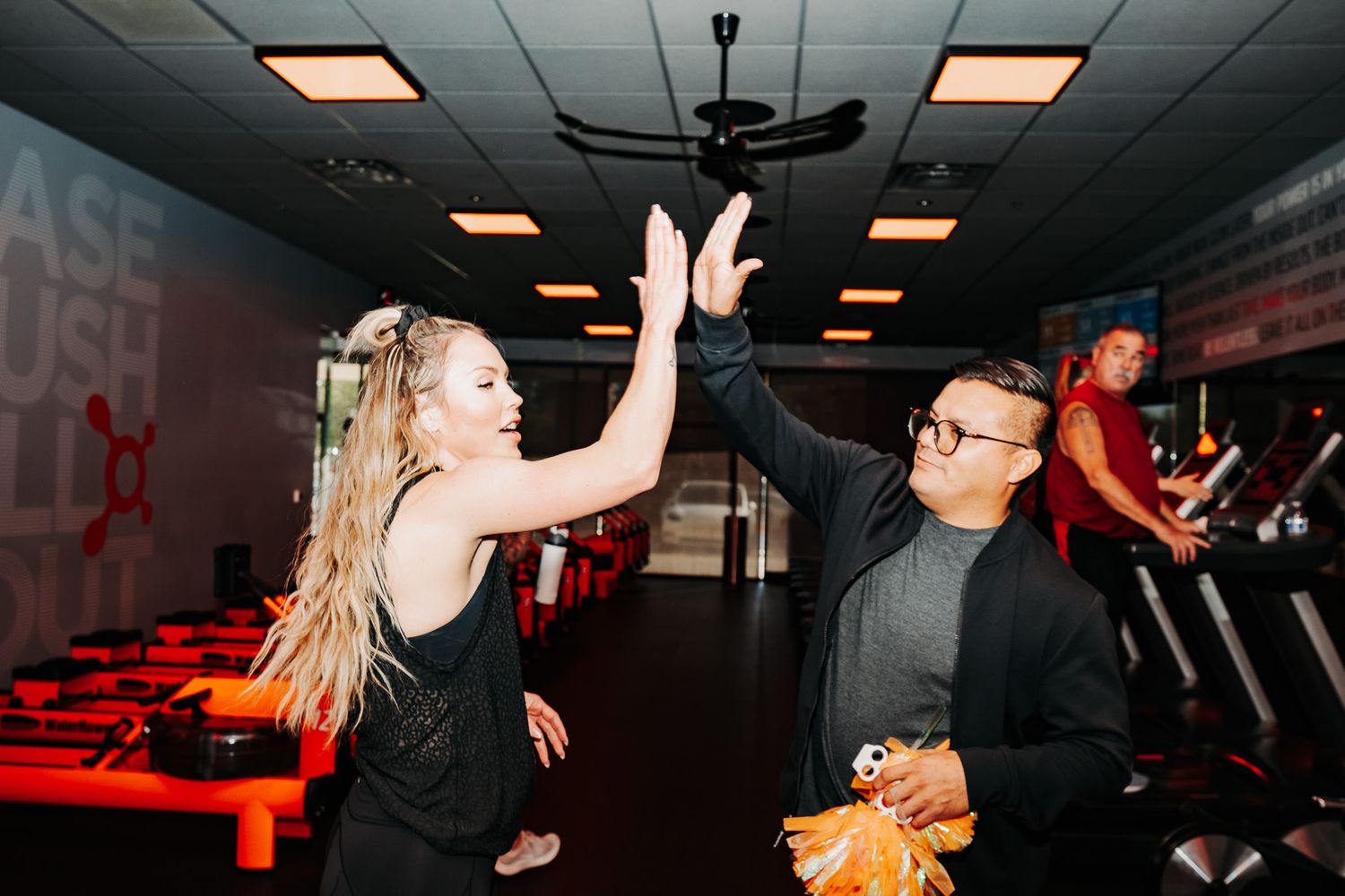 People celebrating and dancing in a dim-lit fitness studio with orange lighting and workout equipment.
