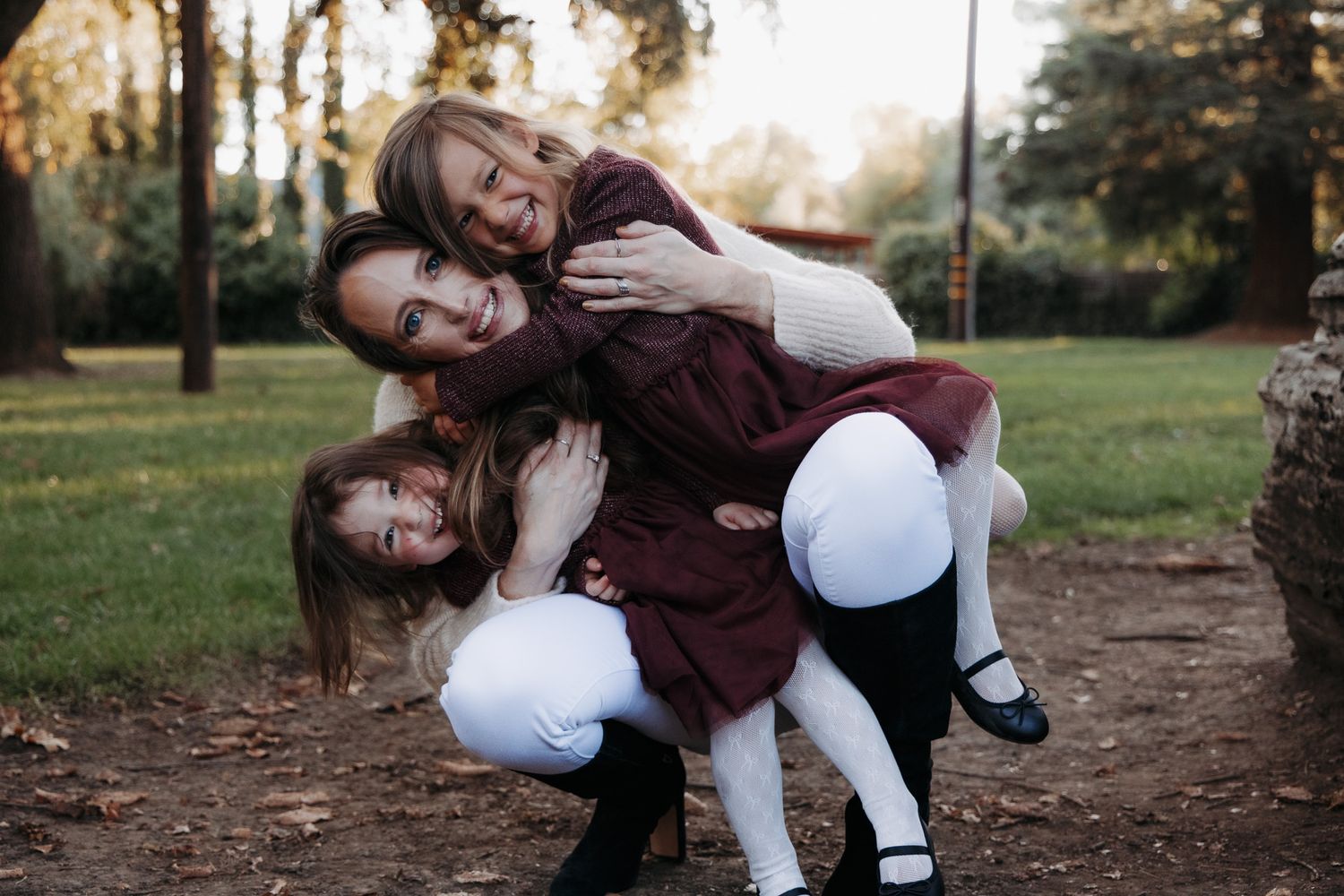 A person in a burgundy sweater and white pants sits playfully on a dirt path in an autumn setting.