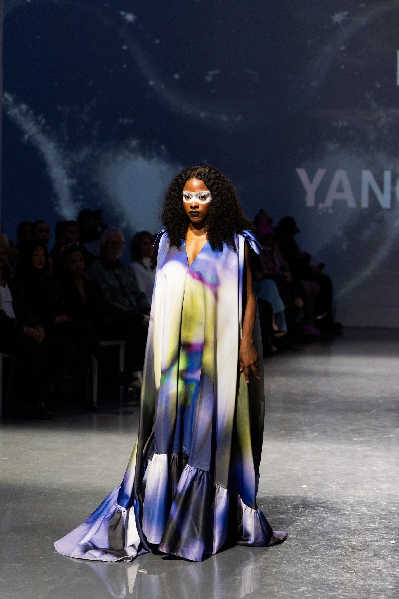 Flowing iridescent cape dress floats dramatically on fashion runway under night sky.