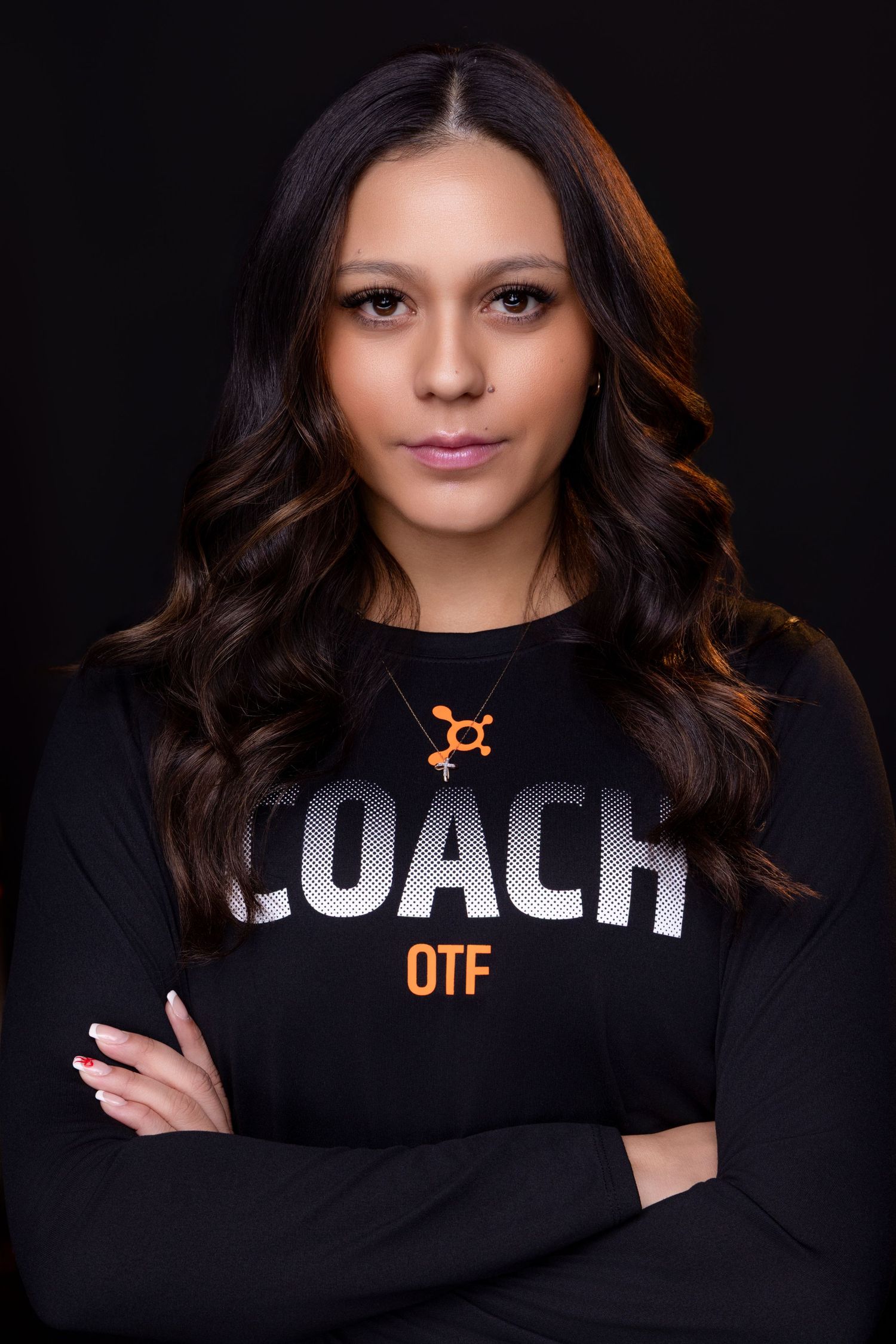 Fitness coach wearing black long sleeve shirt with orange OTF logo stands confidently with arms crossed against dark background.