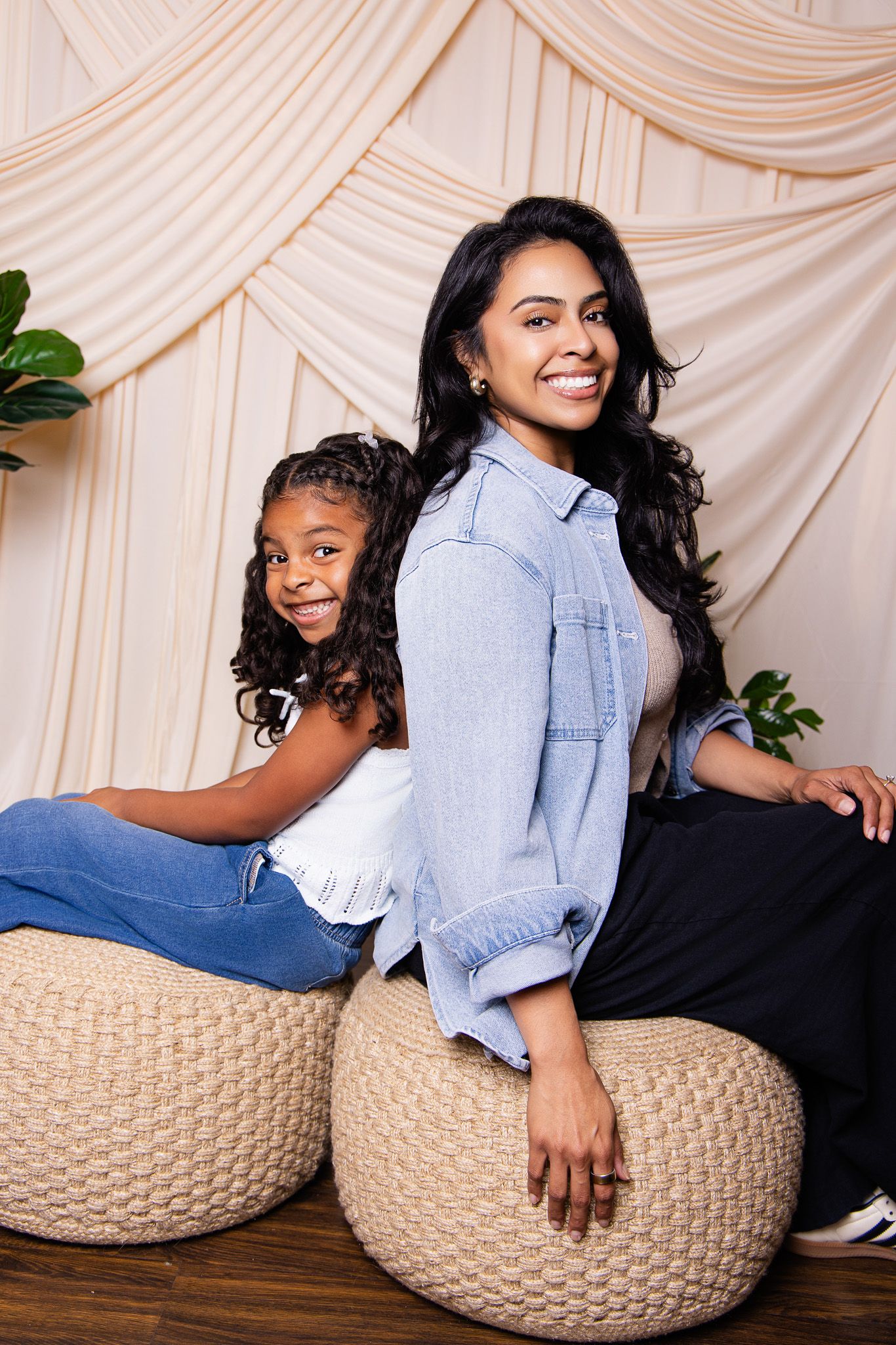 A woman and young girl smile while sitting back-to-back on woven ottomans in a studio with draped backdrop.