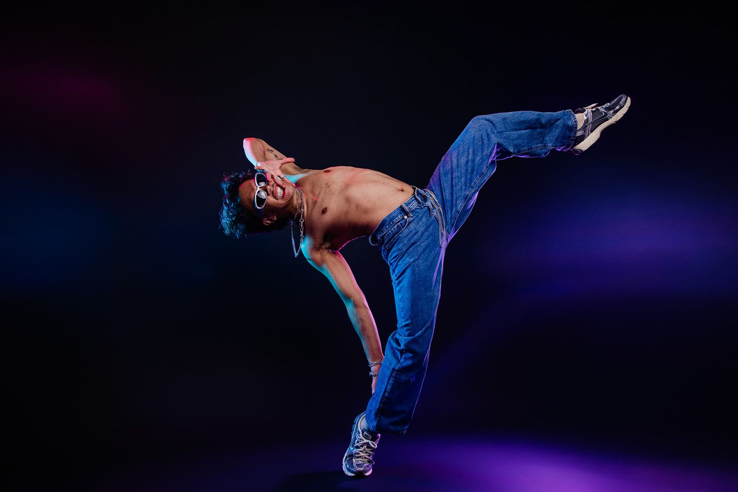Shirtless male dancer performing dynamic breakdance move in blue jeans against dark studio background with purple lighting.