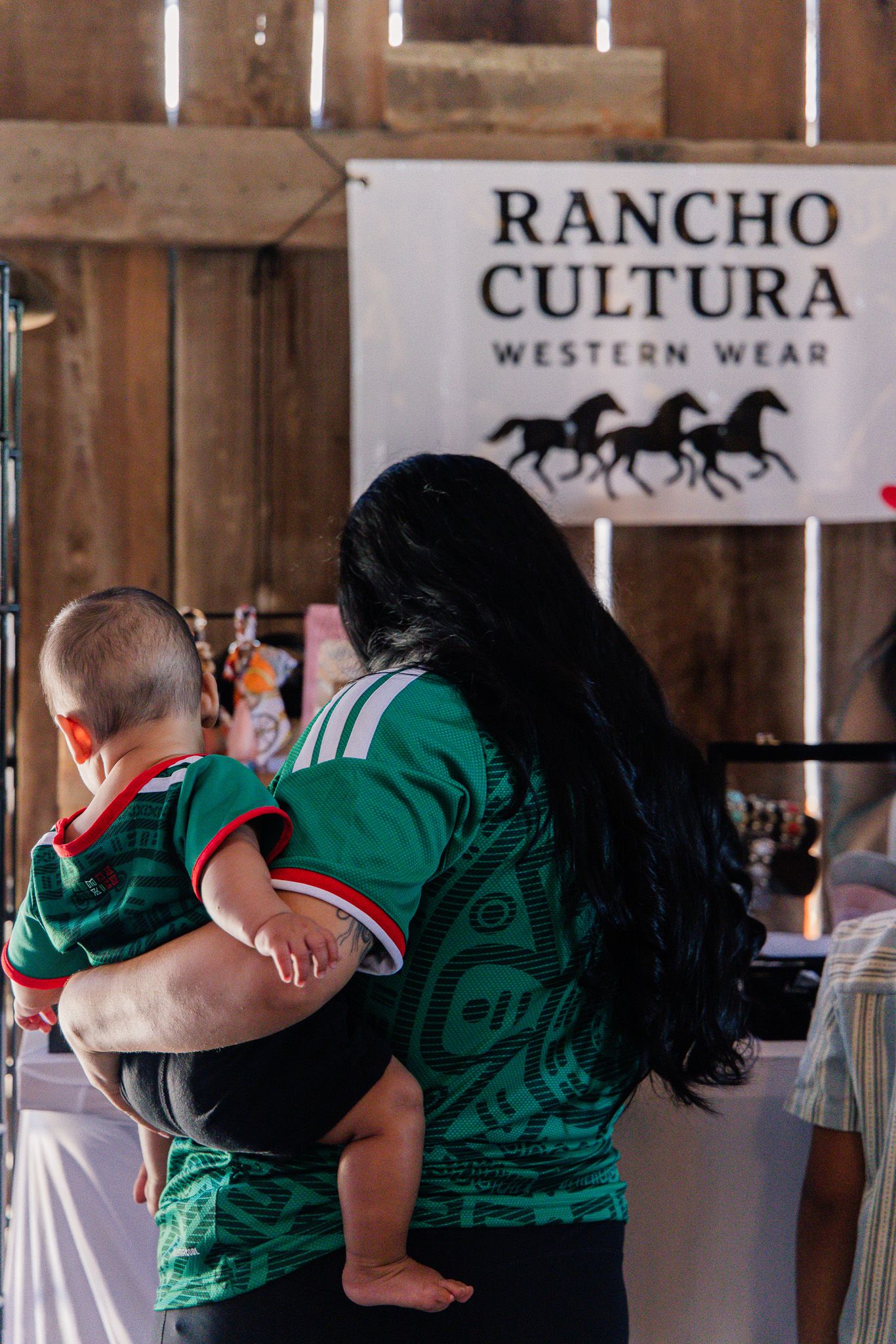 Woman with long black hair holding a baby in a green jersey near Rancho Cultura Western Wear sign.