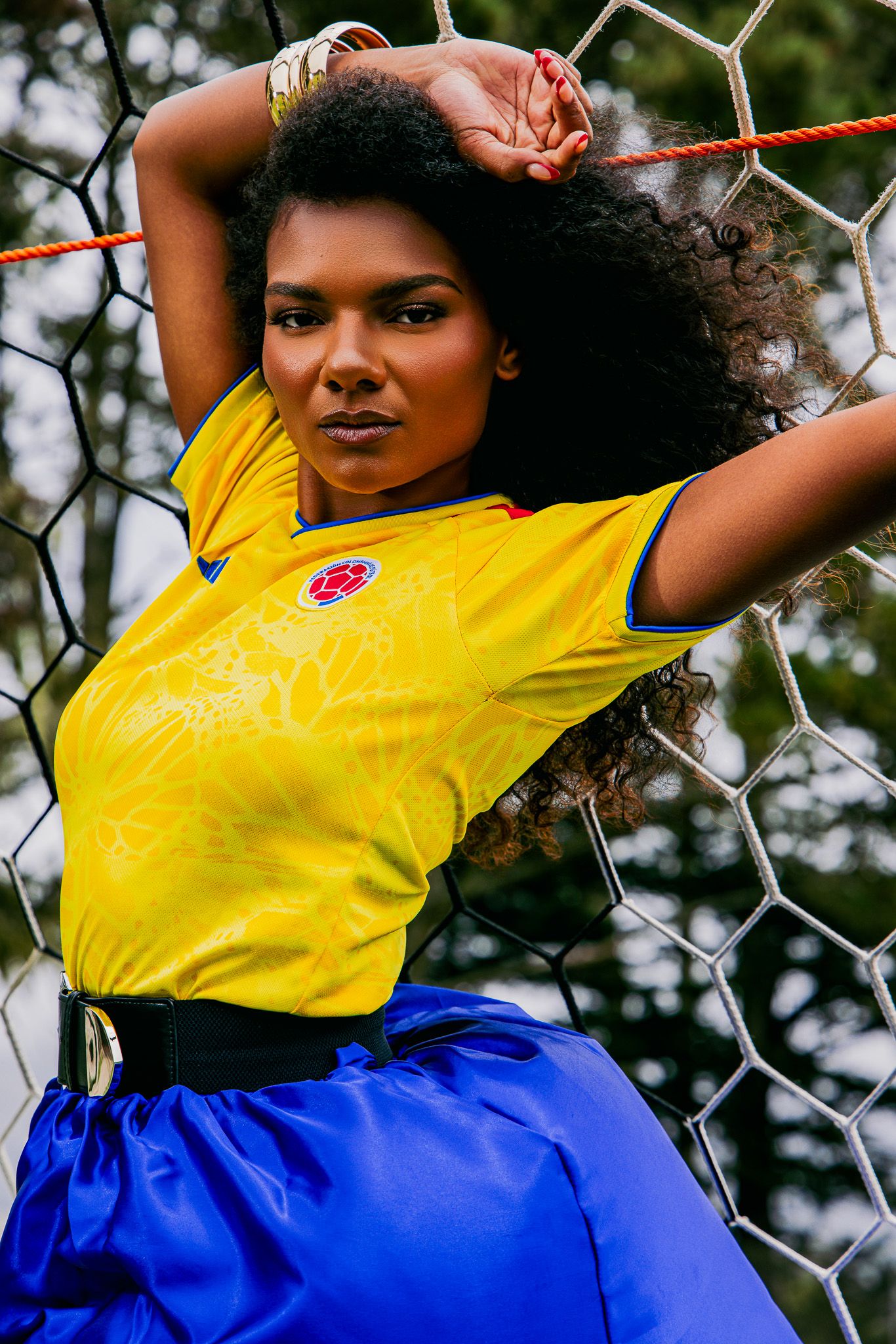 Woman in yellow Colombia soccer jersey and blue skirt poses against a football goal net outdoors.