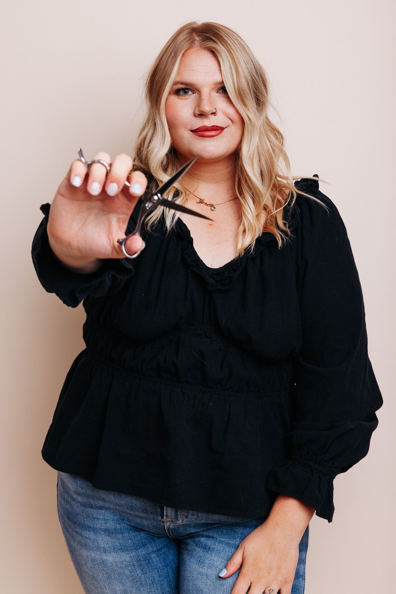 Blonde woman in black blouse holding hair scissors toward camera with confident expression.