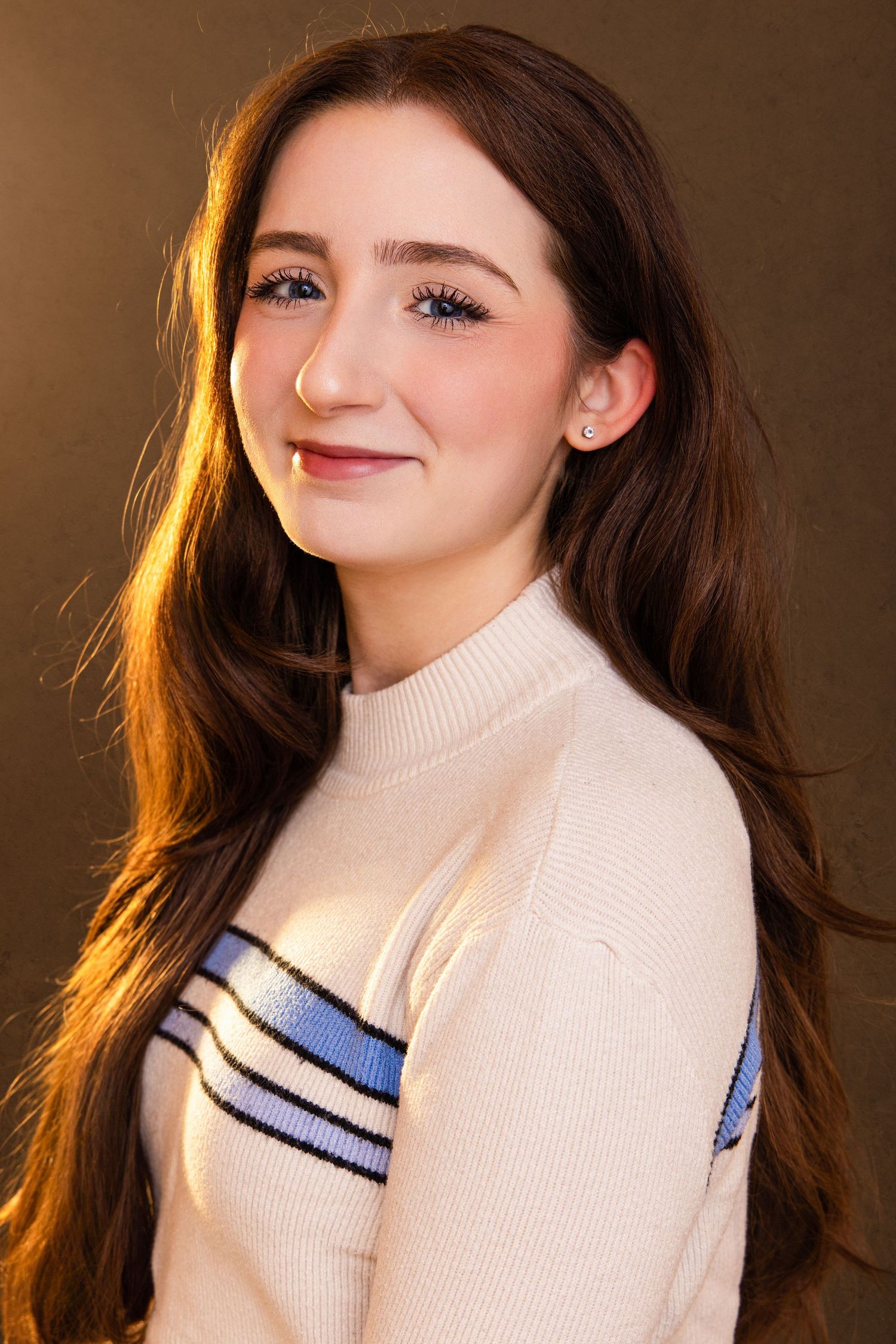 Young woman with long brown hair smiling, wearing a cream striped turtleneck sweater against a warm brown background.