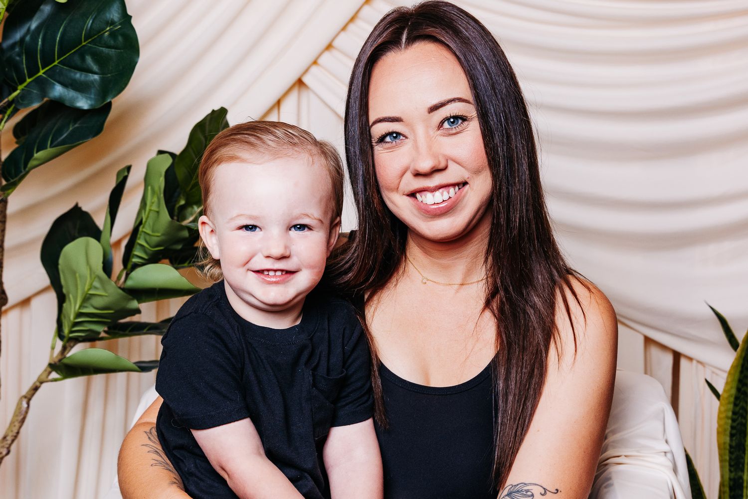 A smiling woman with long dark hair holds a happy red-haired toddler in a black shirt against a light backdrop.