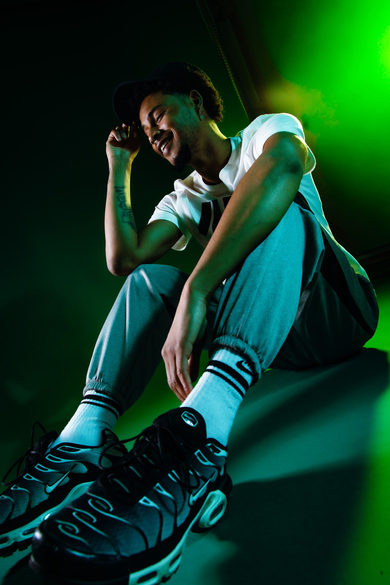 Young man in cap, white tee, joggers and Nike TN sneakers laughing under green studio light.