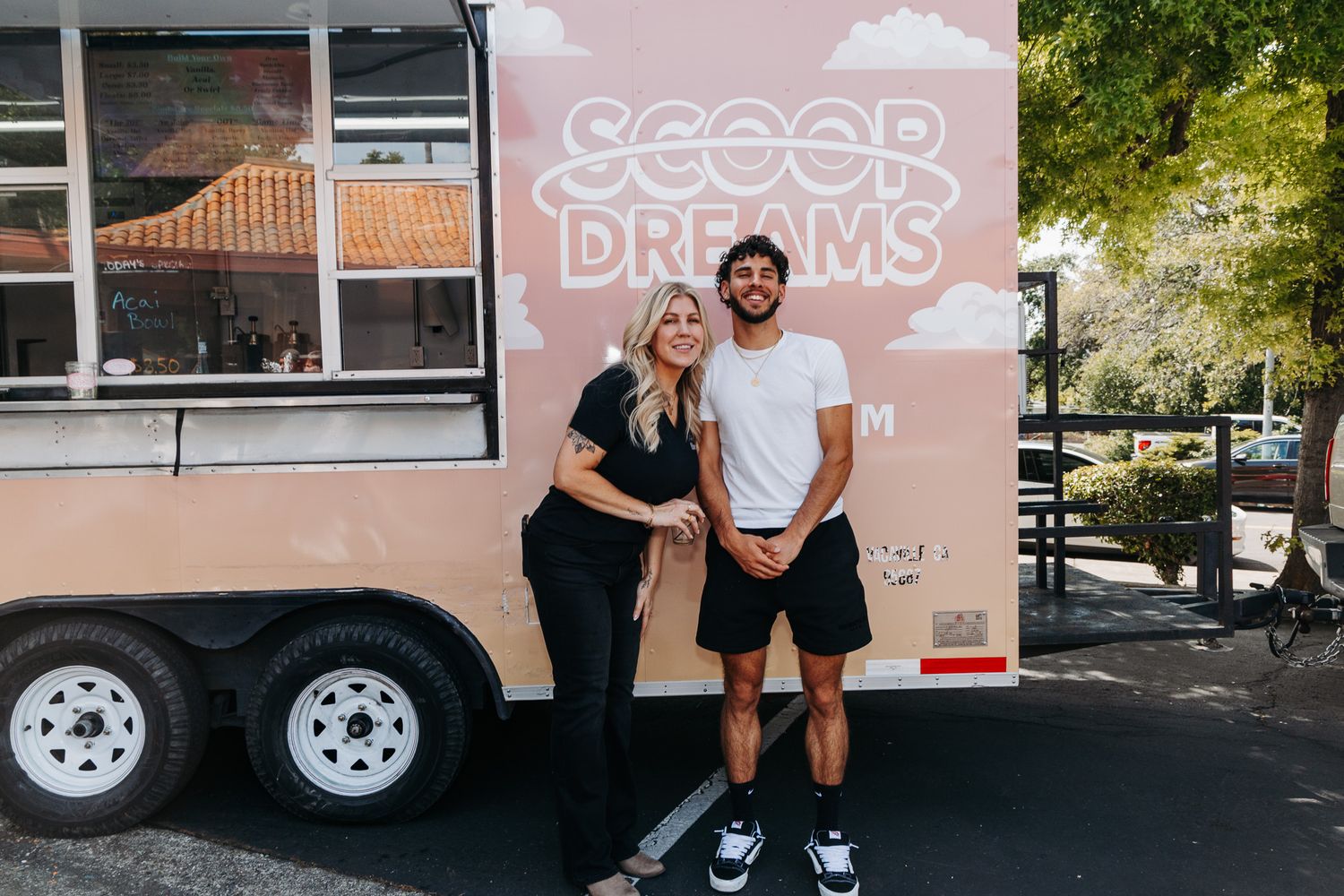 Couple stands together in front of pink Scoop Dreams ice cream trailer at outdoor event.