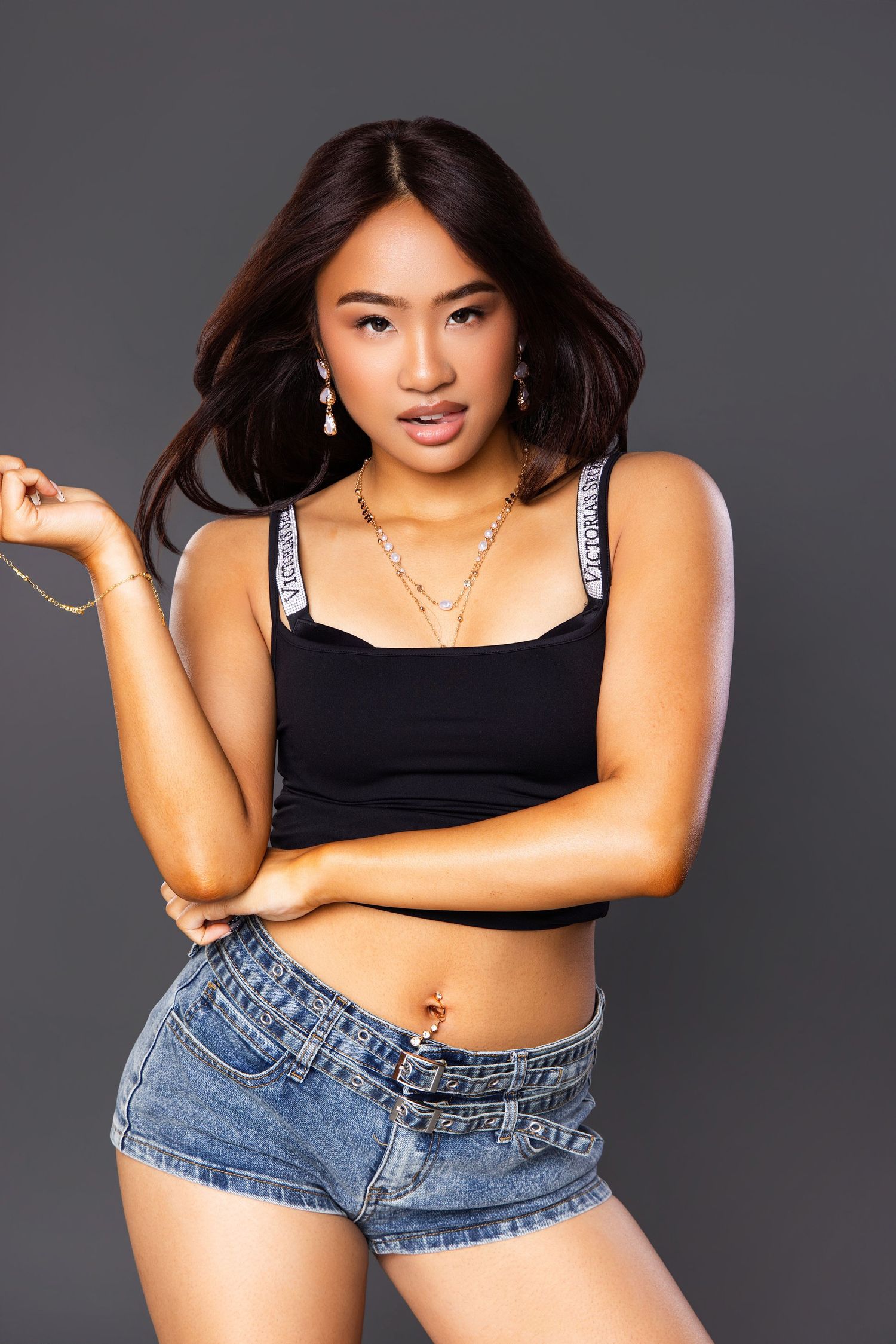Young woman in black crop top and denim shorts posing confidently in studio with gray background.
