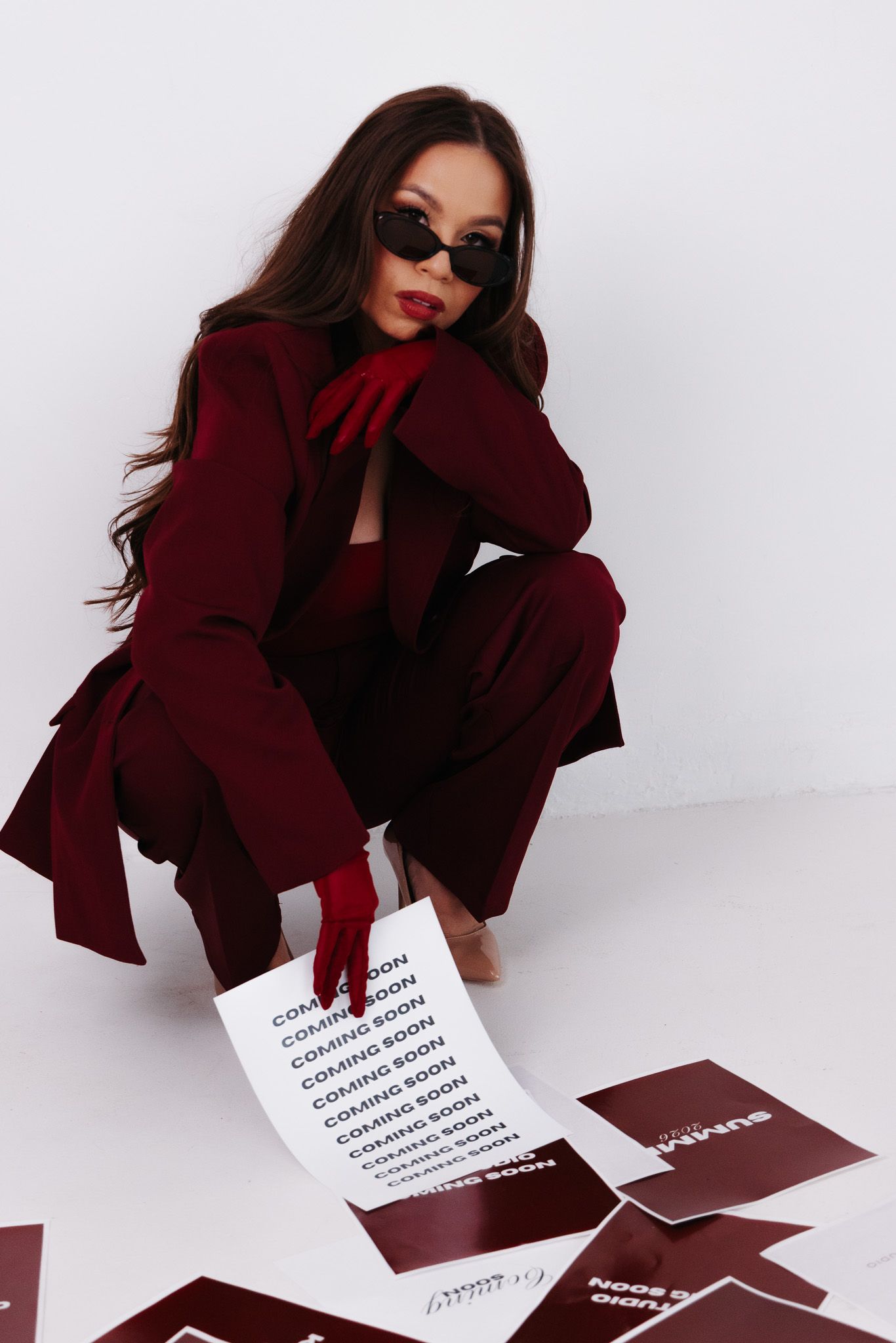 Woman in burgundy suit and sunglasses crouching with coming soon papers on white studio floor.