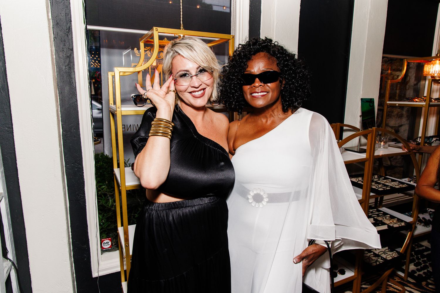 Two women pose together at an event, one in black and one in white dress, both wearing gold accessories.