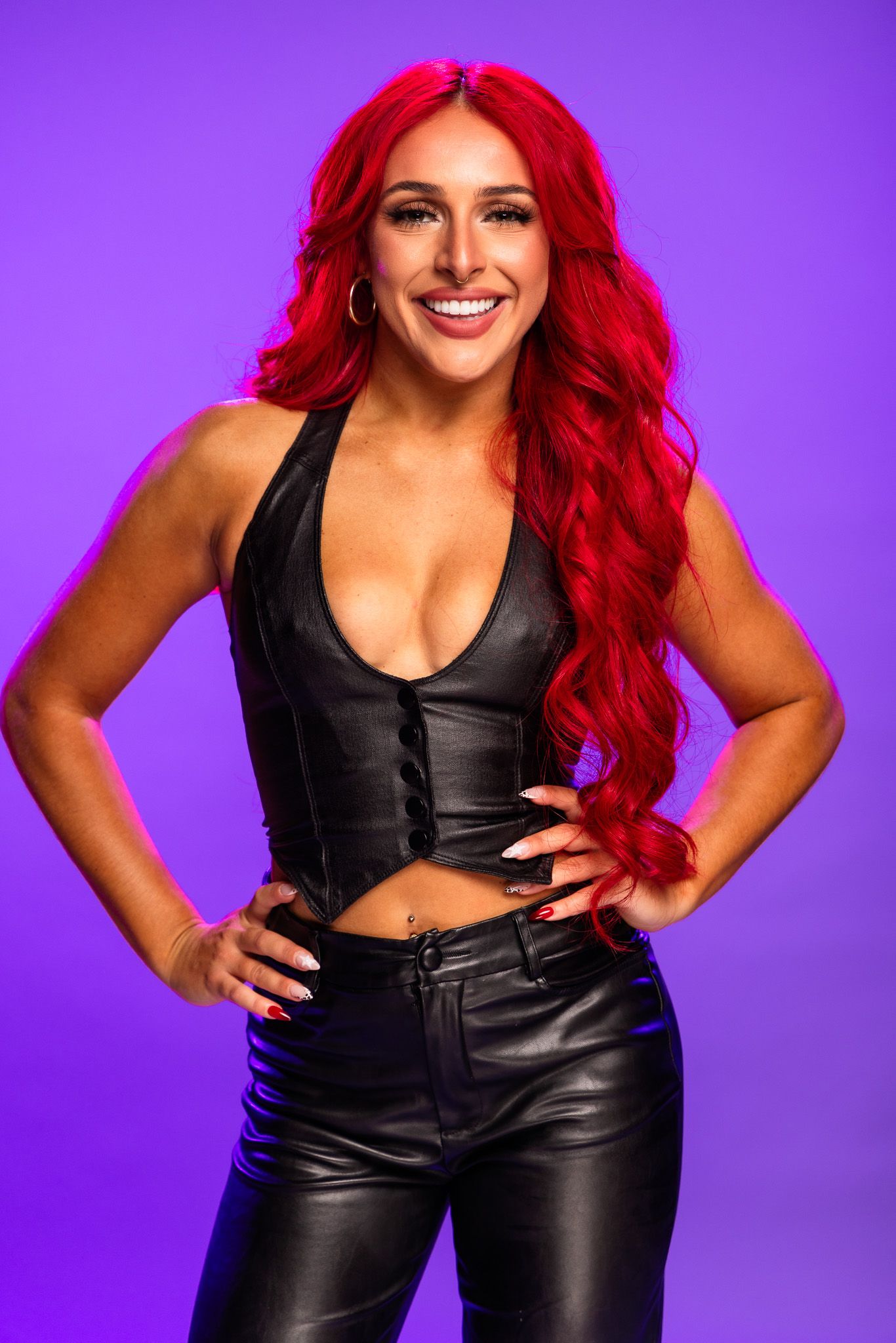 Woman with vibrant red hair wearing black leather outfit poses confidently against a purple background.