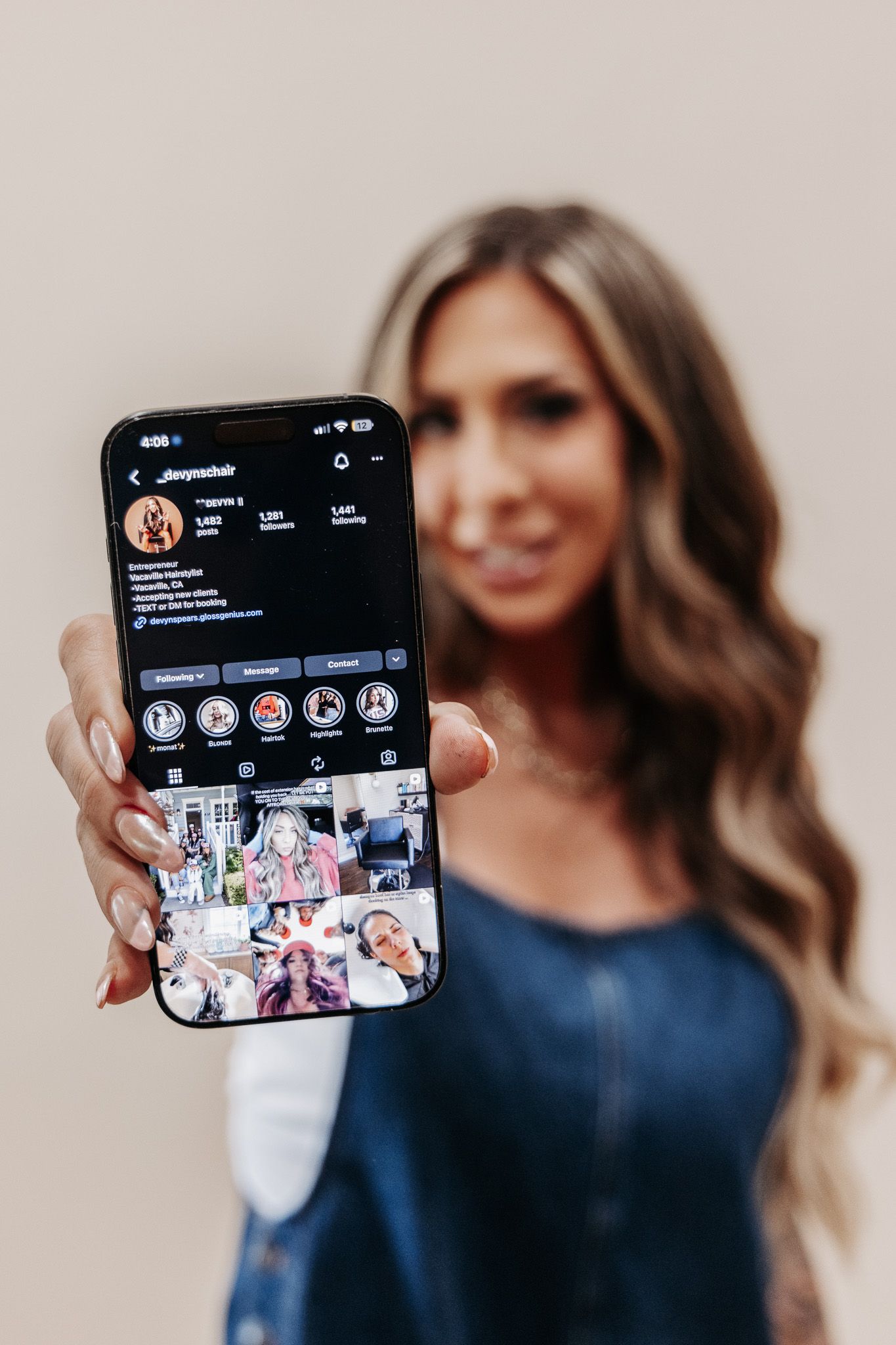 Blonde woman holding smartphone displaying Instagram profile page in dark mode, wearing denim top.