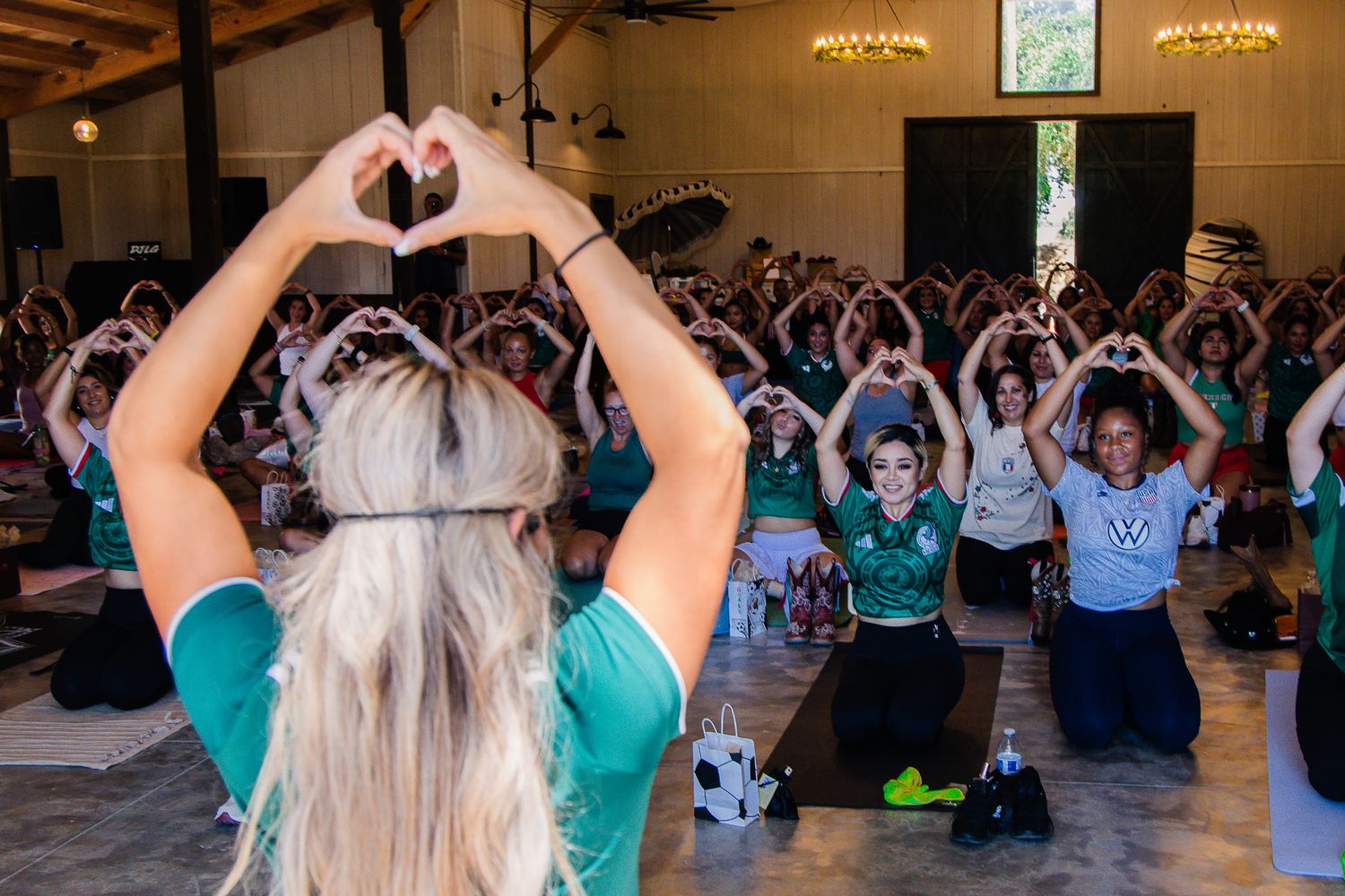 Group yoga class with participants making heart shapes with their hands, led by a blonde instructor in a green shirt.
