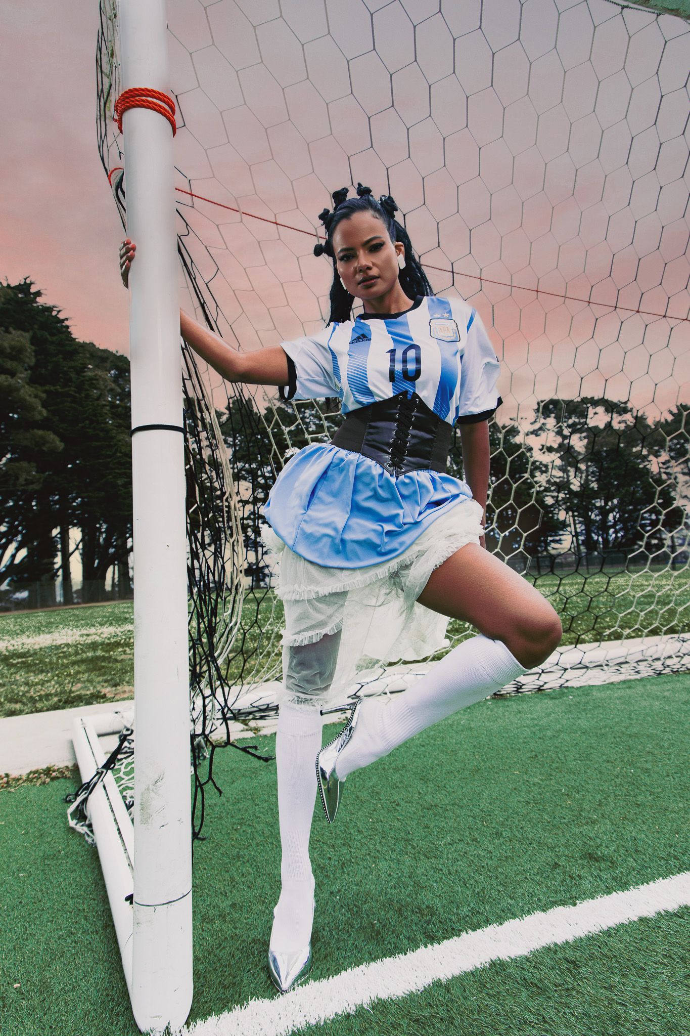 Woman in Argentina soccer jersey, corset, blue skirt, and white boots posing at a football goal post on green turf.