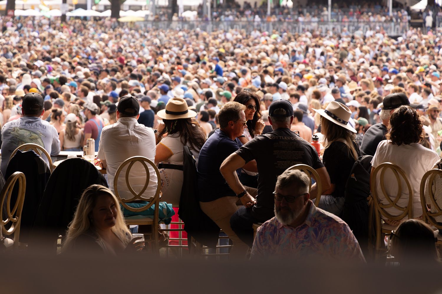 Large outdoor festival crowd wearing hats seated under bright sunlight at a summer music event.