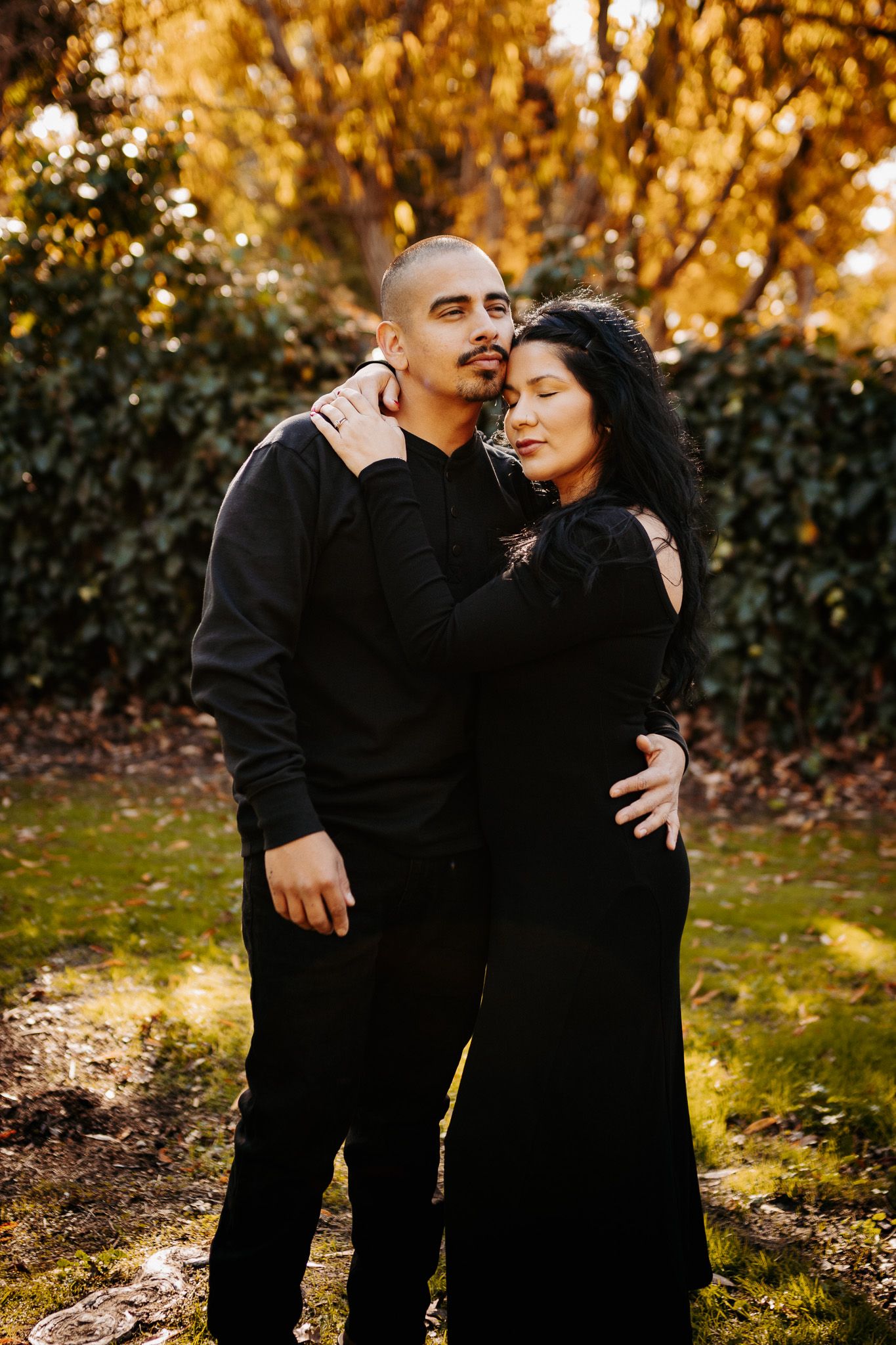 A couple wearing black outfits embrace tenderly in a park with golden autumn foliage in the background.