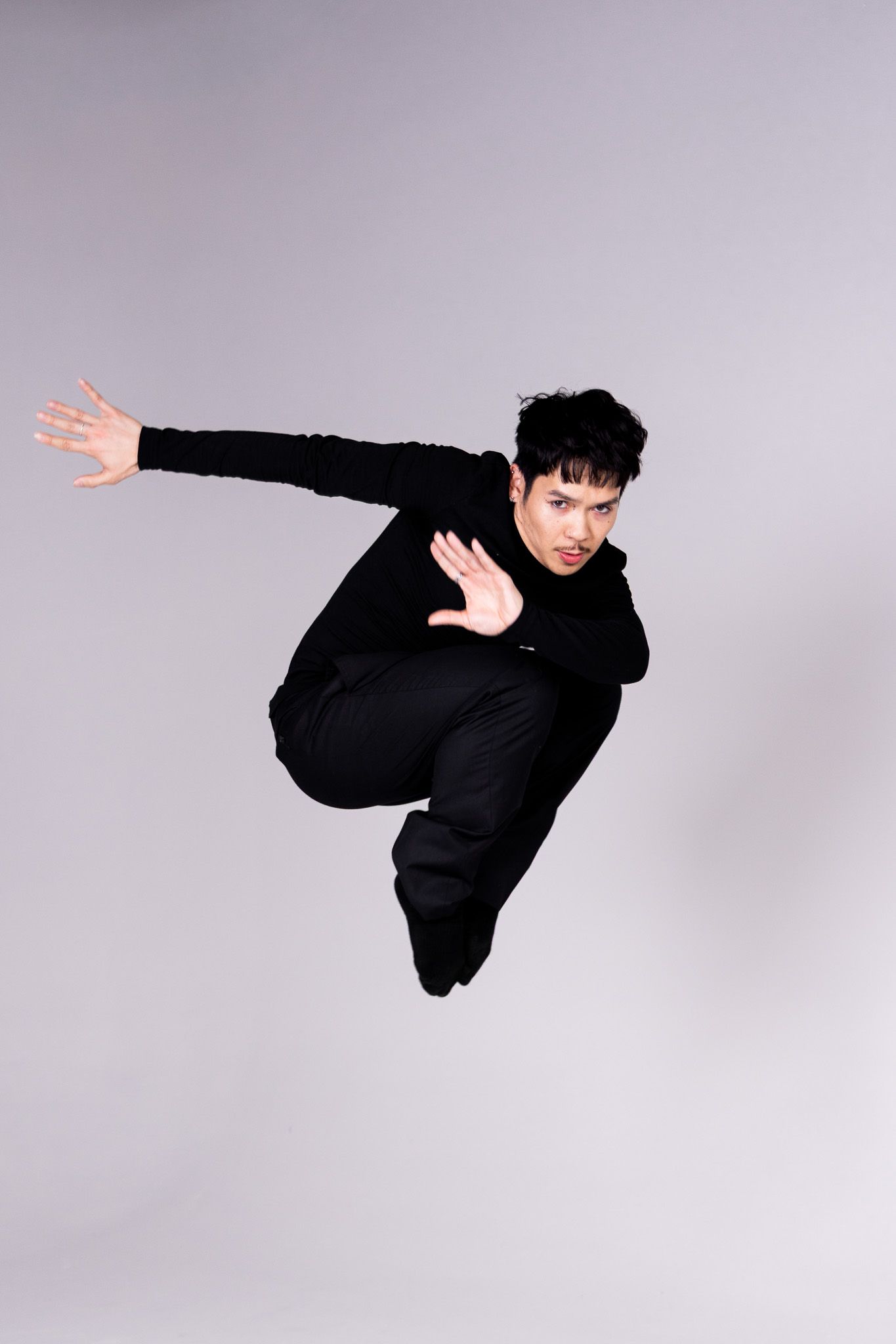 Dynamic mid-air jump pose of performer in black outfit against studio backdrop.