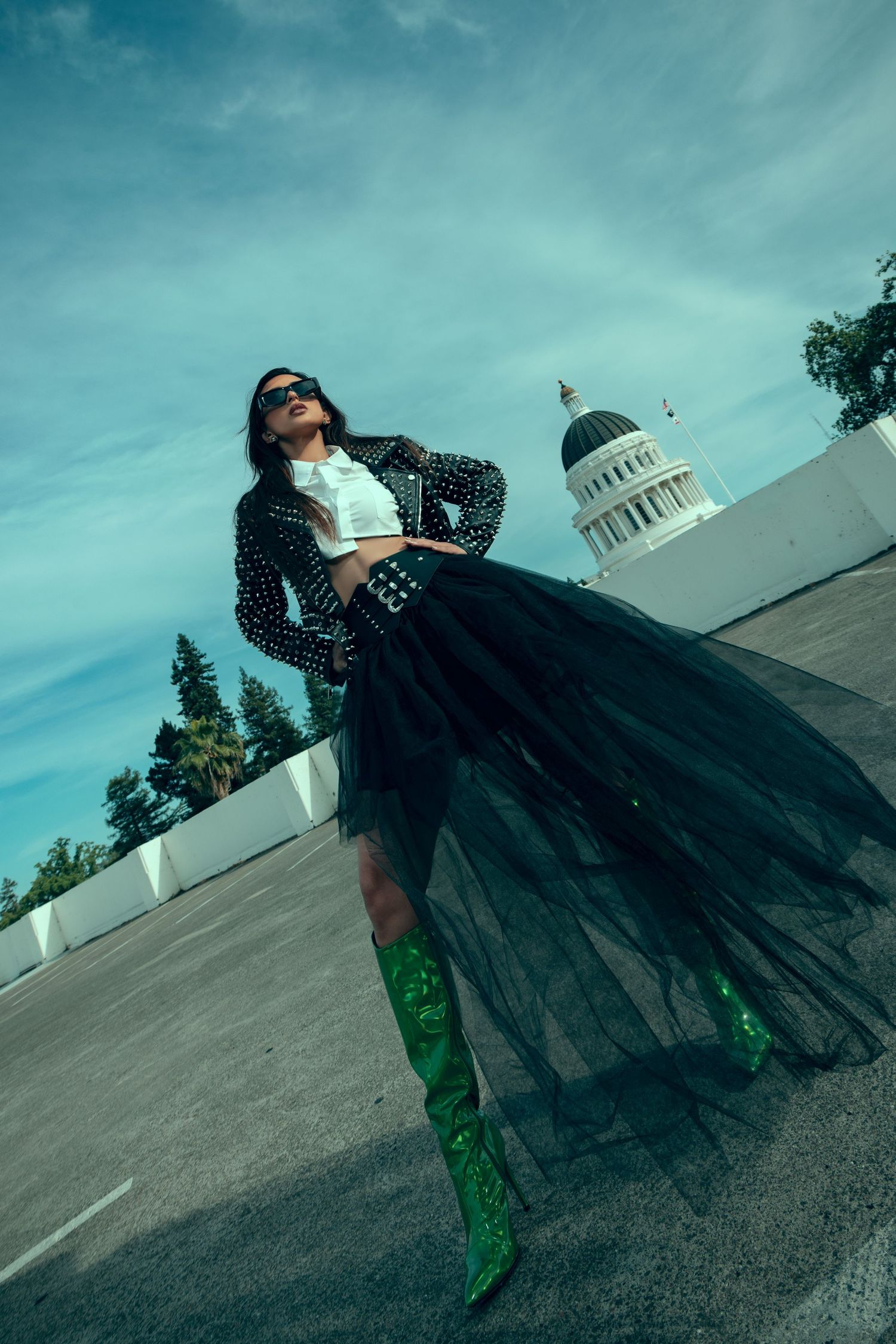A figure in green pants and dramatic black flowing fabric poses dynamically against a blue sky with white architecture.