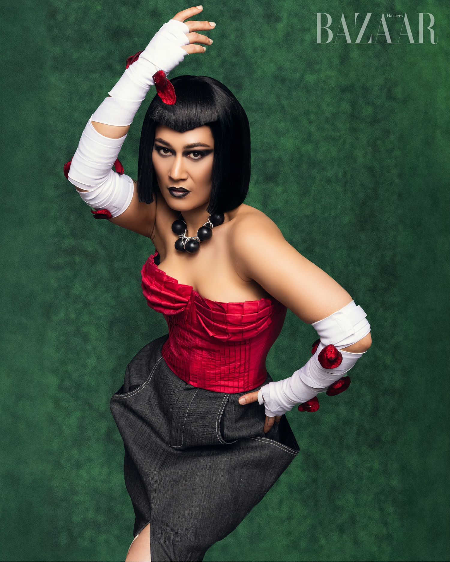 Fashion model in red and black dress with white gloves poses dramatically against emerald green backdrop.