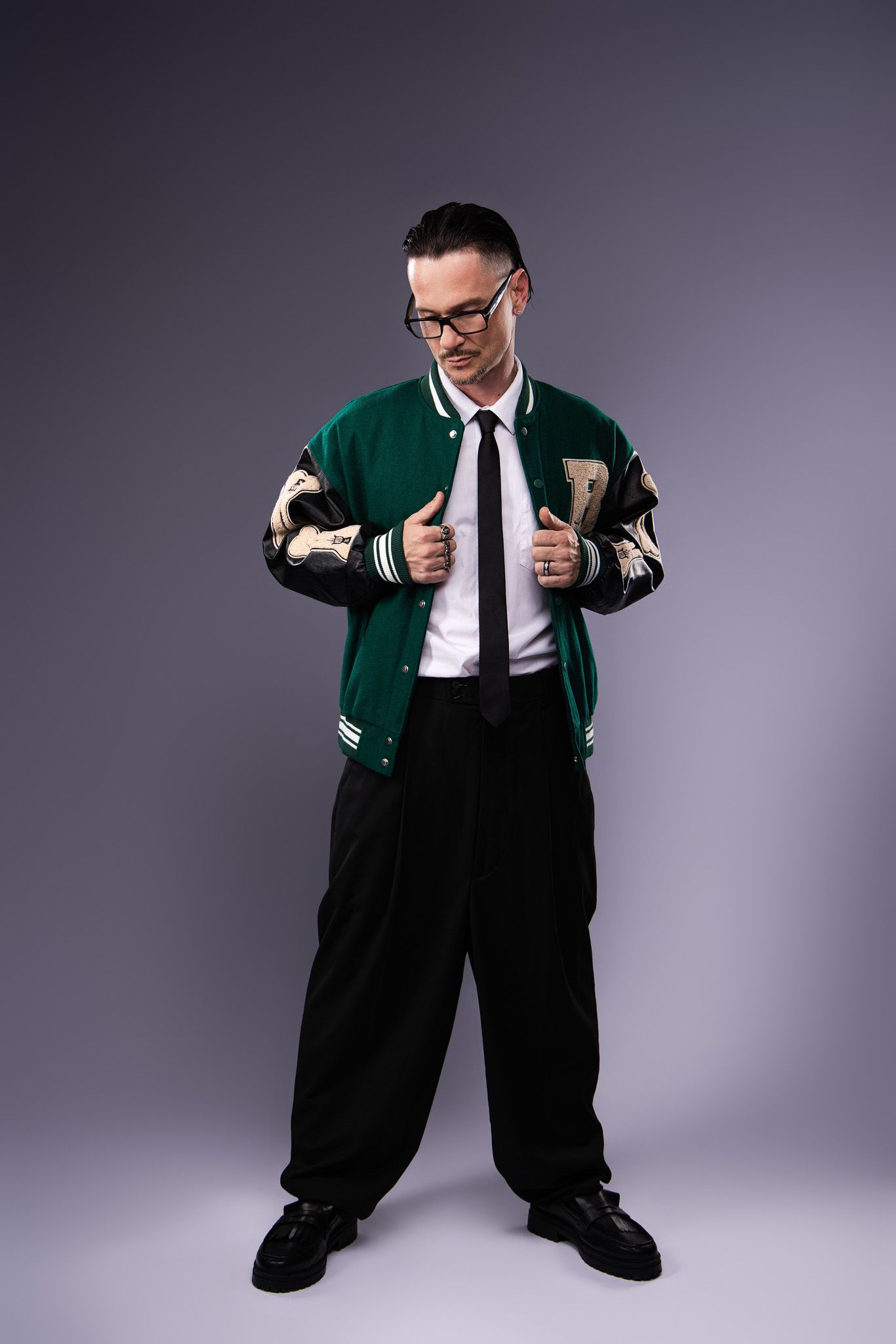 Person wearing oversized black pants and green varsity jacket strikes dynamic poses against gray studio background.