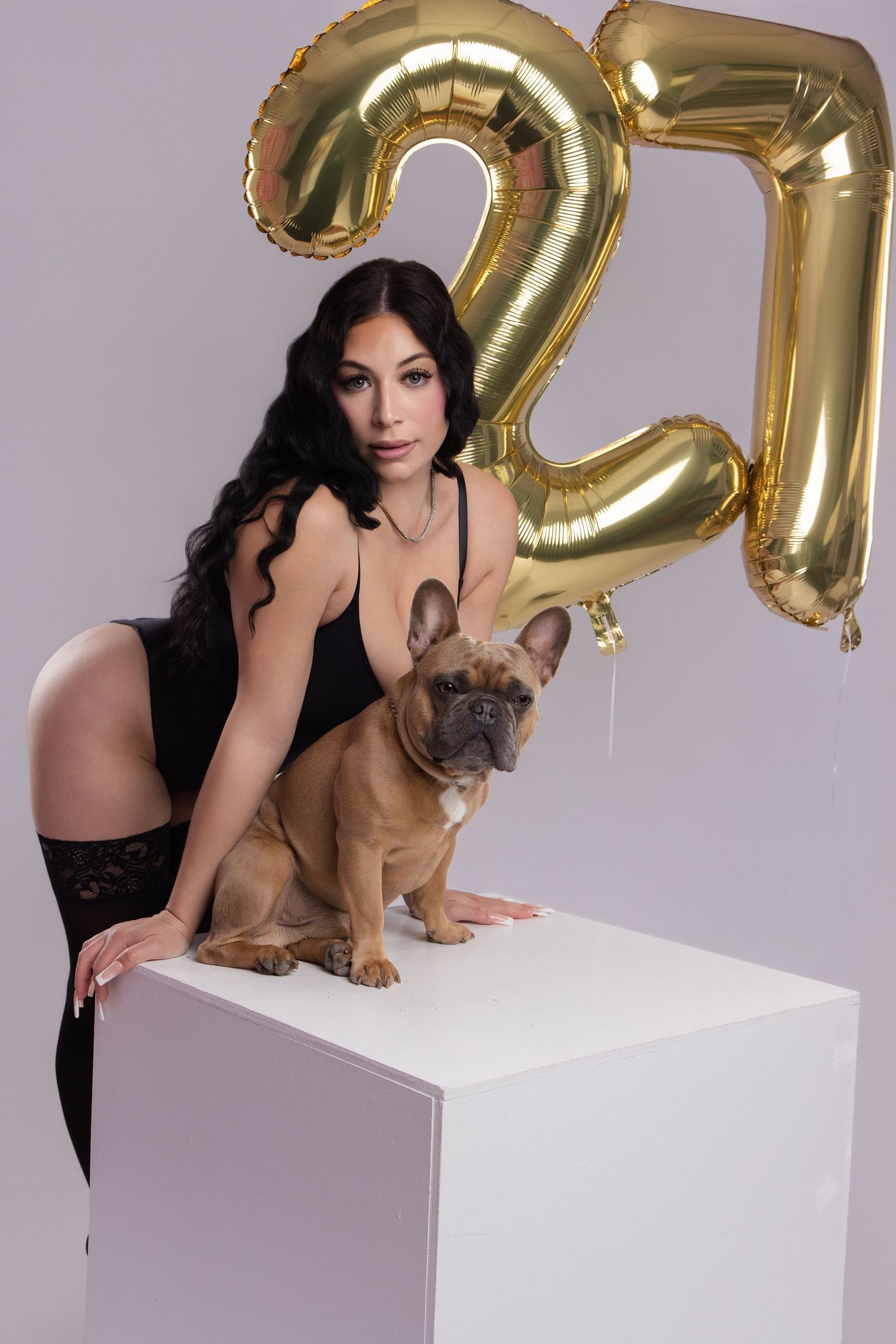 A woman in black dress poses with a French Bulldog on a white pedestal with gold balloon numbers in the background.
