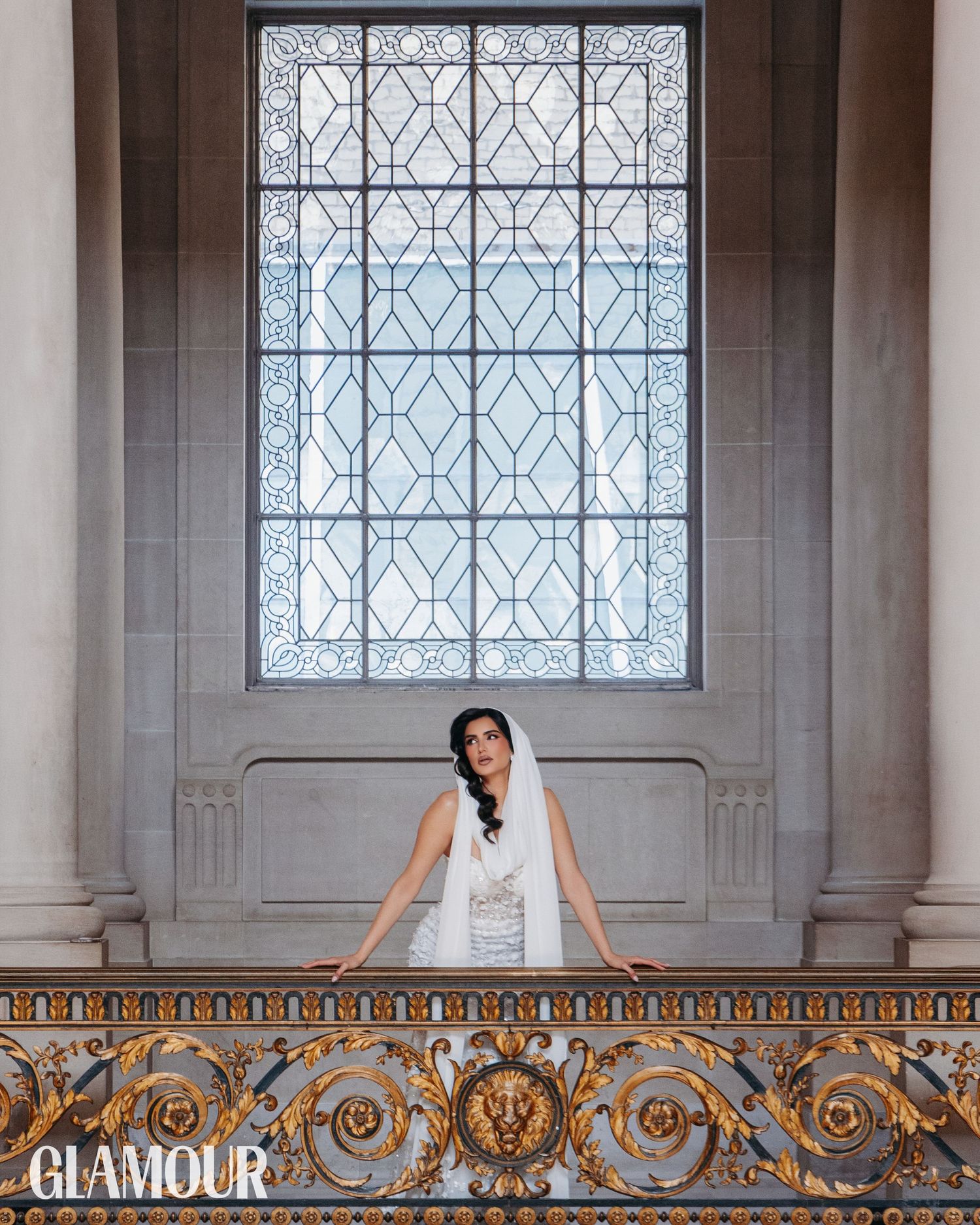 Elegant bride in white dress poses on ornate gold-trimmed balcony against decorative window in classic architectural setting.