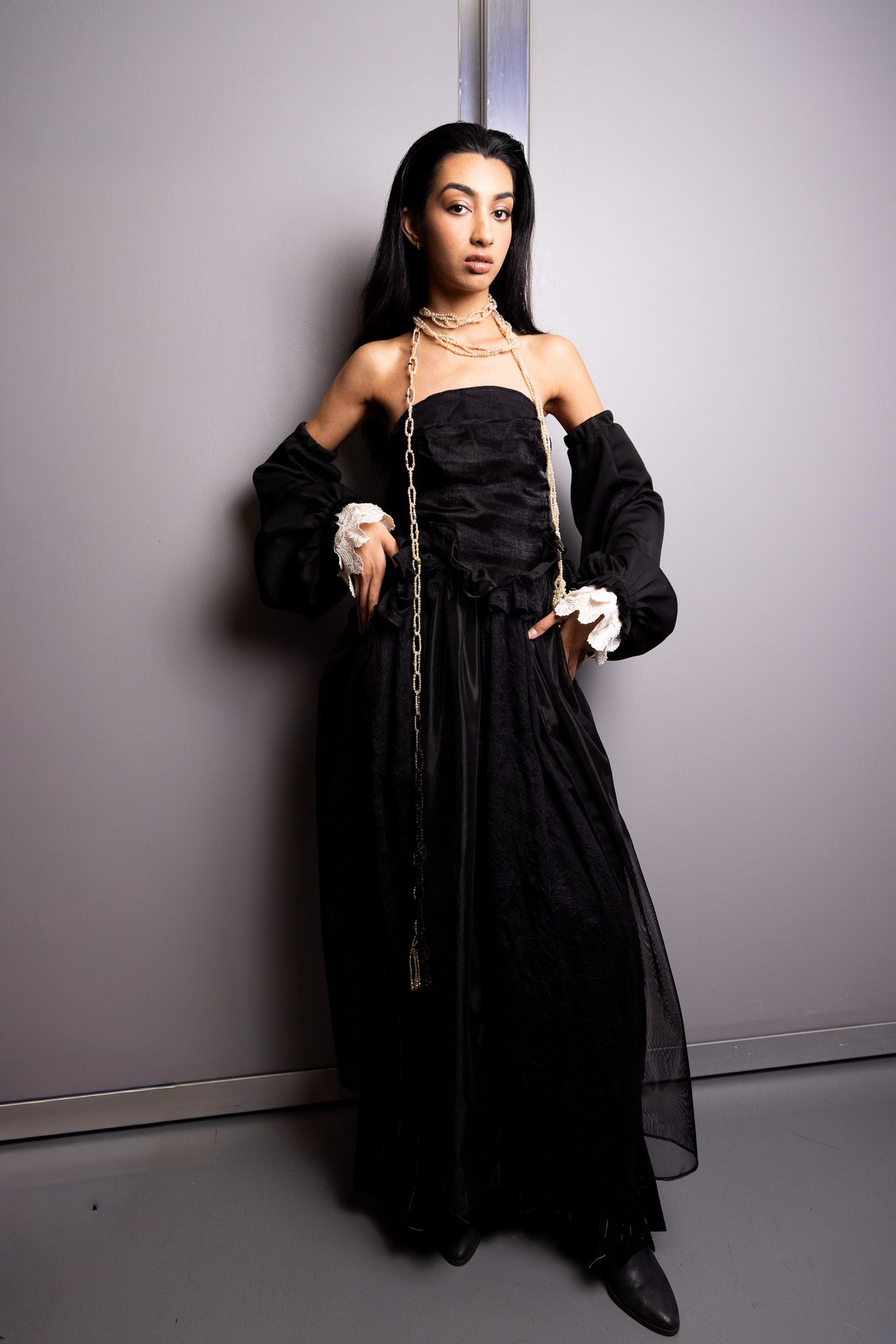 Model in dramatic black off-shoulder gown with sheer details and delicate chain accessories poses against grey wall.