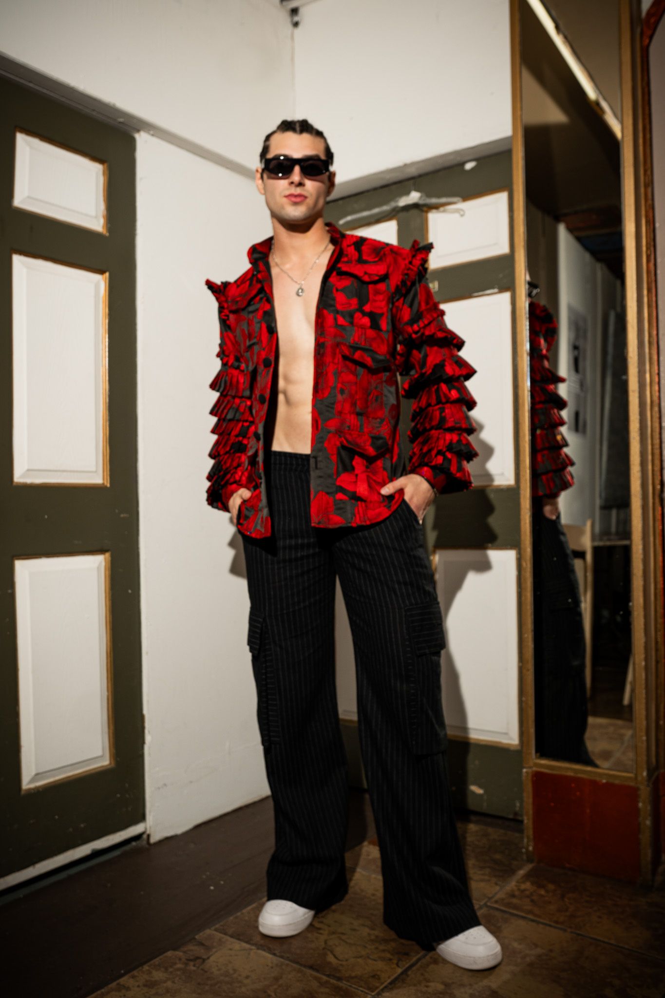 A person poses in a dramatic red and black ruffled jacket with sunglasses in a hallway setting.