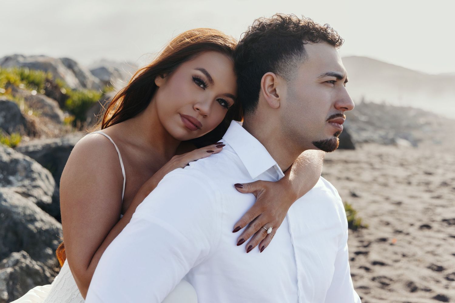 Romantic couple embraces on sandy beach with rocky coastline and misty mountains in background.