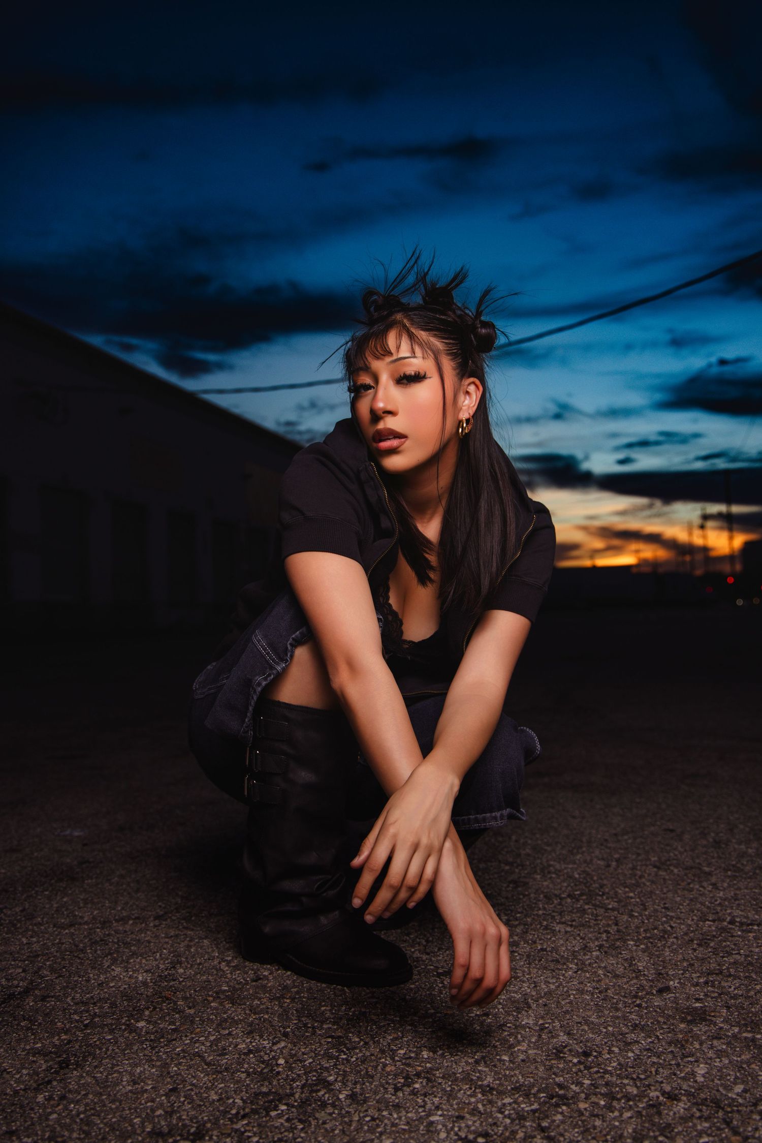 A figure crouches on dark ground against a dramatic sunset sky with power lines visible in the background.