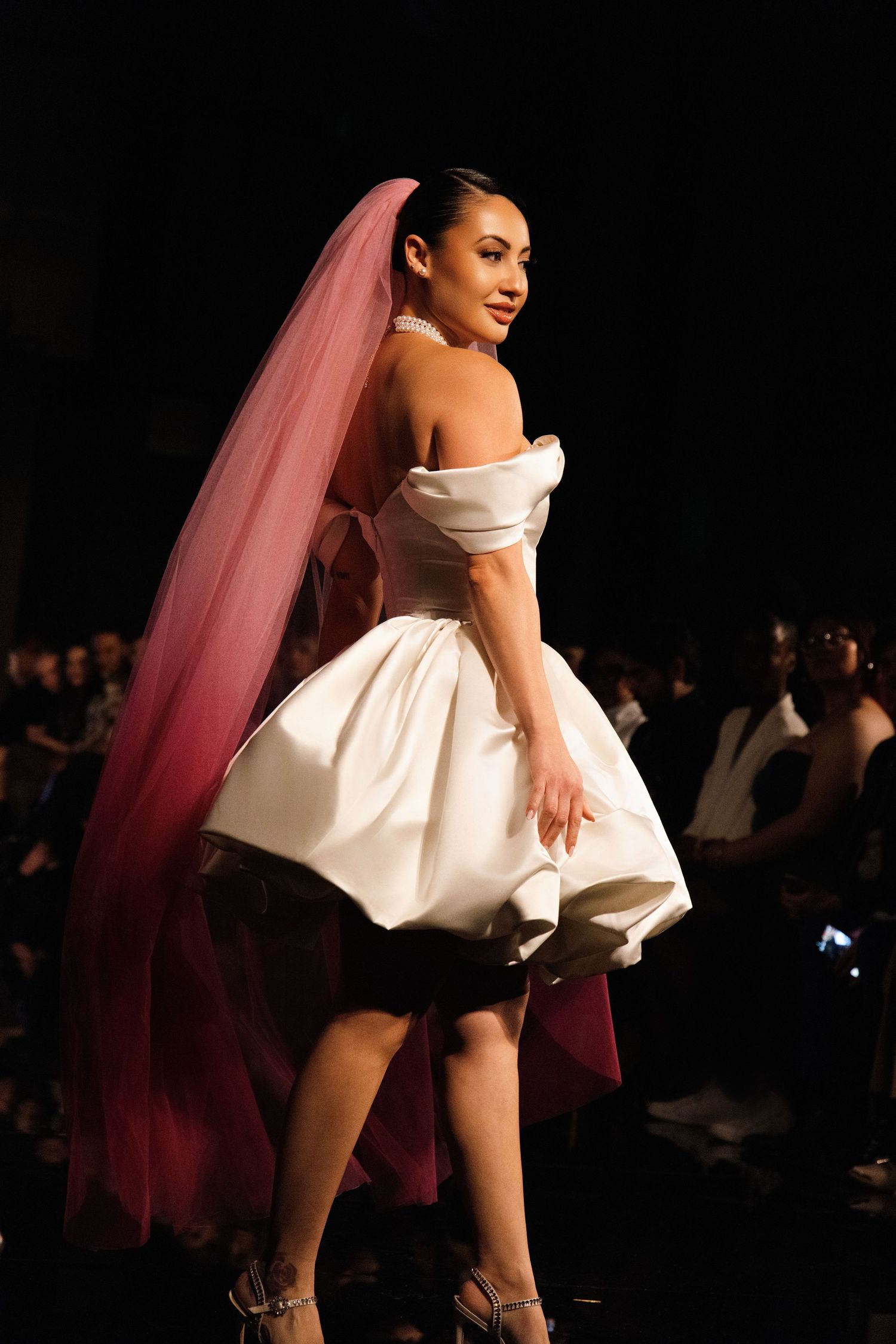 Model showcases a short white bridal dress with flowing veil on a dark fashion runway.