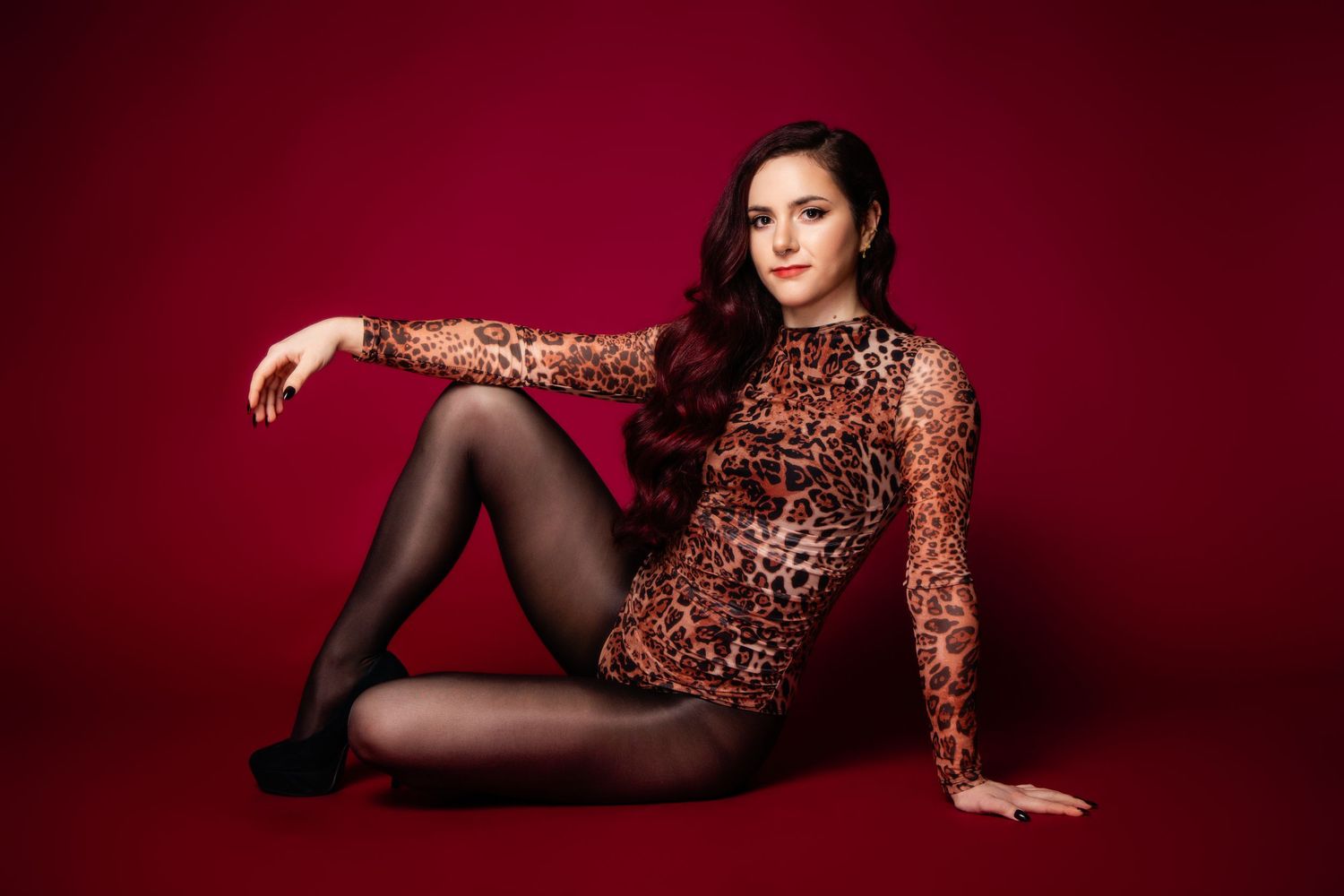 Fashion model in leopard print bodysuit poses against deep red backdrop in glamorous photo series.