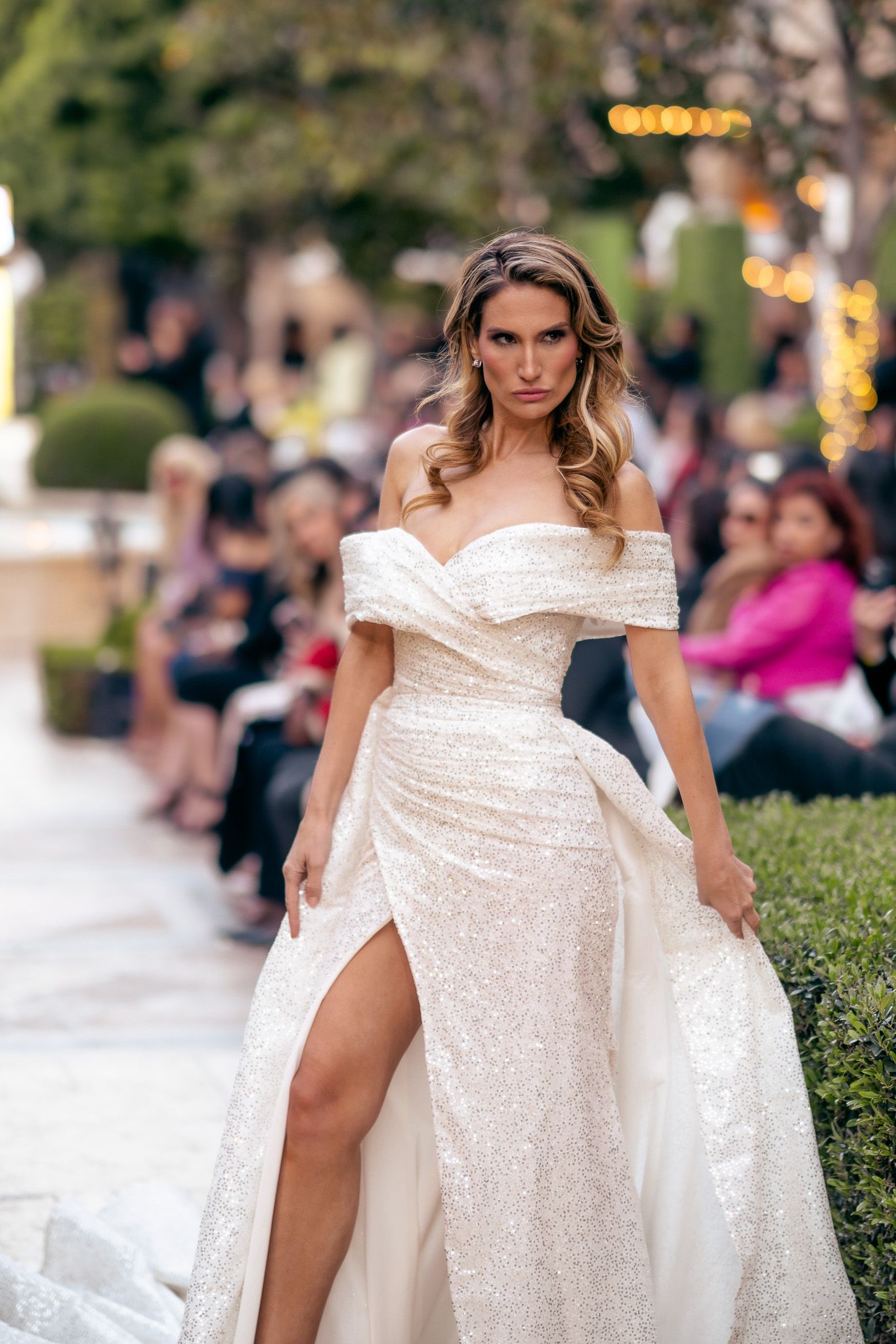 Model walks runway in sparkly off-shoulder white evening gown with thigh-high slit at outdoor fashion show.